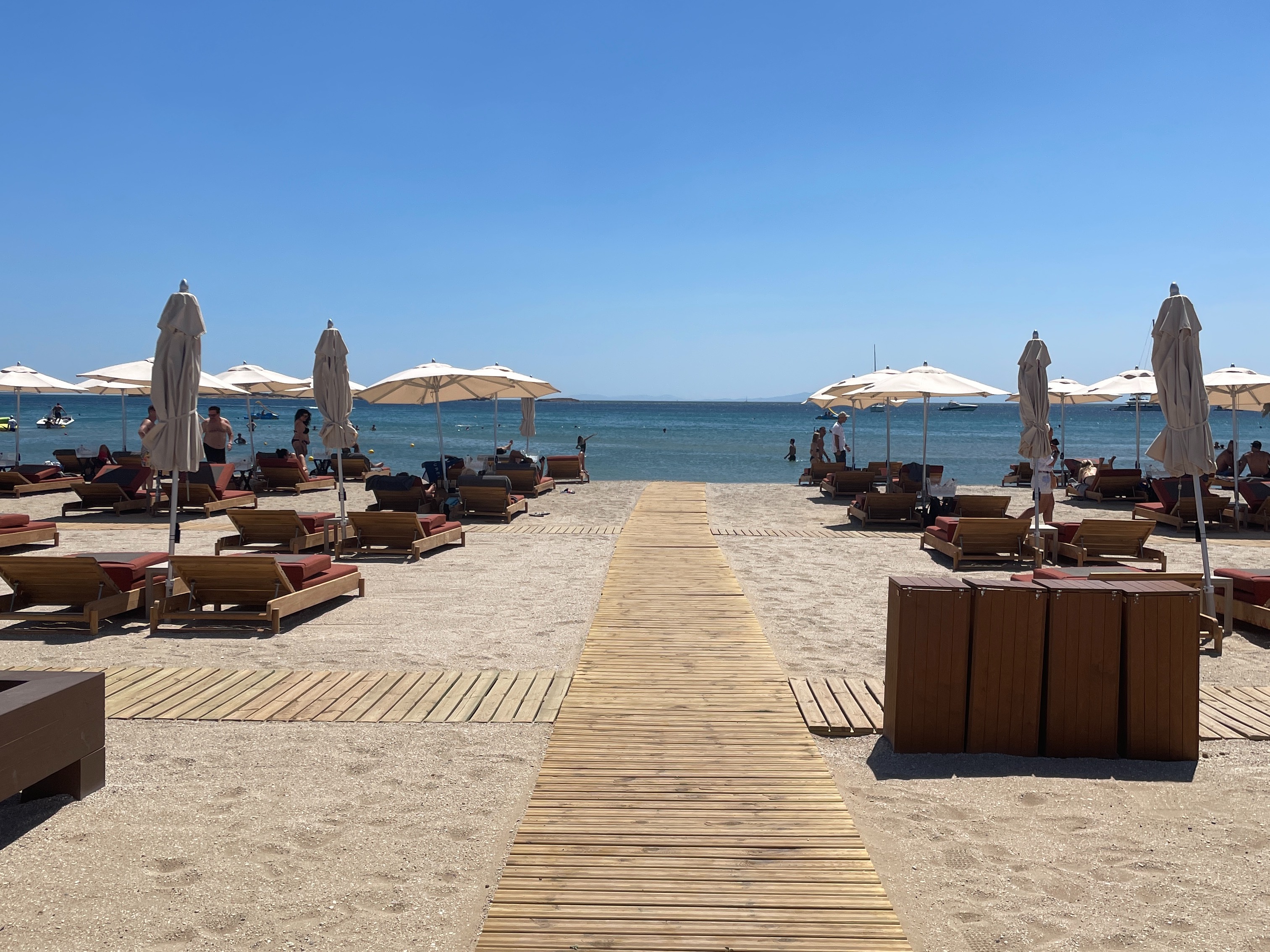 A serene beach scene with a wooden pathway leading to the sea, flanked by sun loungers and umbrellas under a clear blue sky.