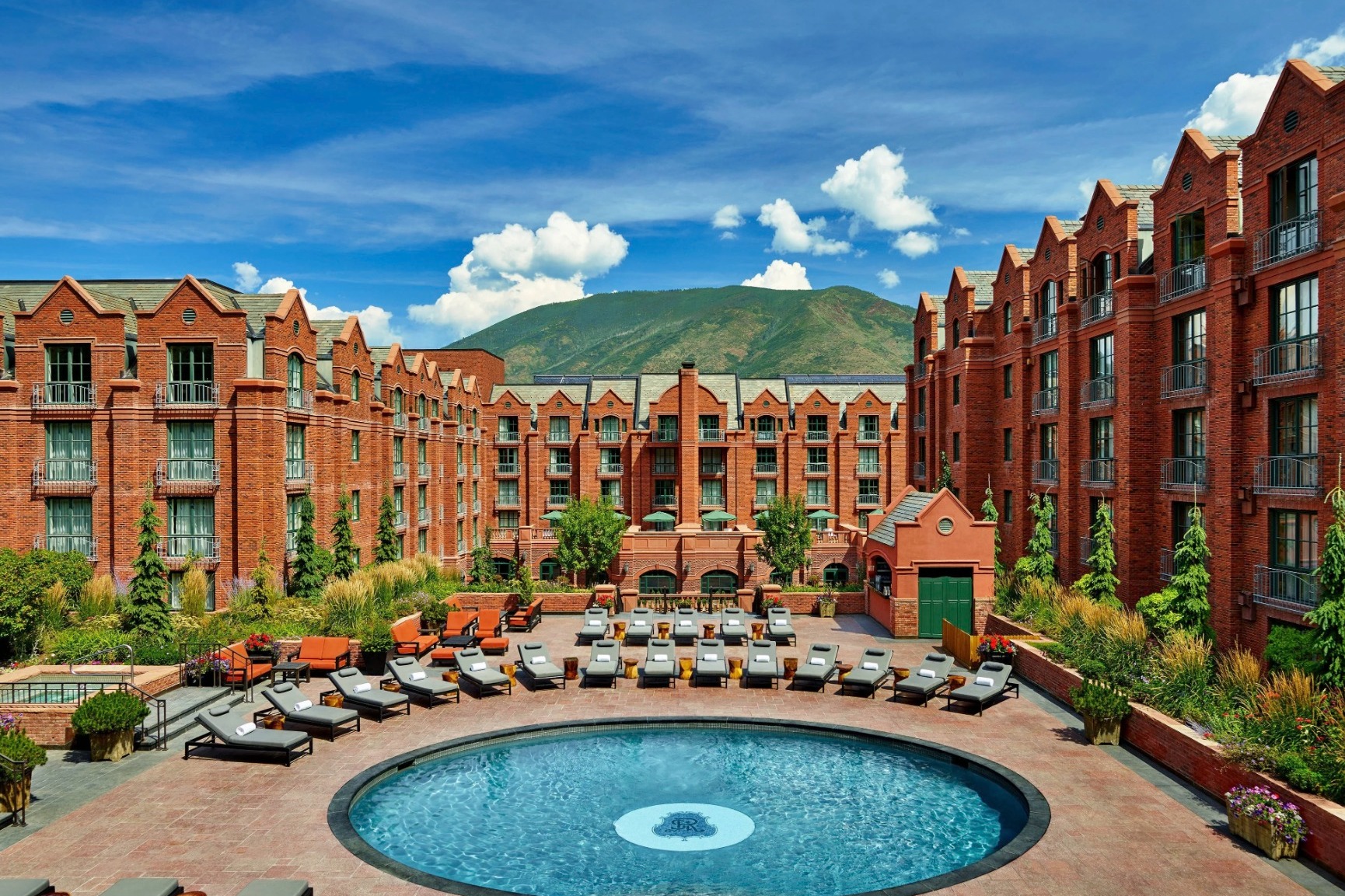 pool surrounded by a brick hotel with blue sky and a green mountain in the background