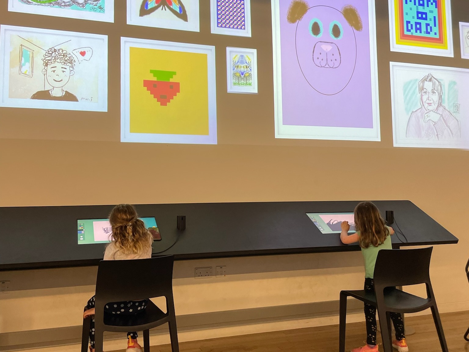 Children interact with digital screens displaying colorful drawings in a creative or educational setting.