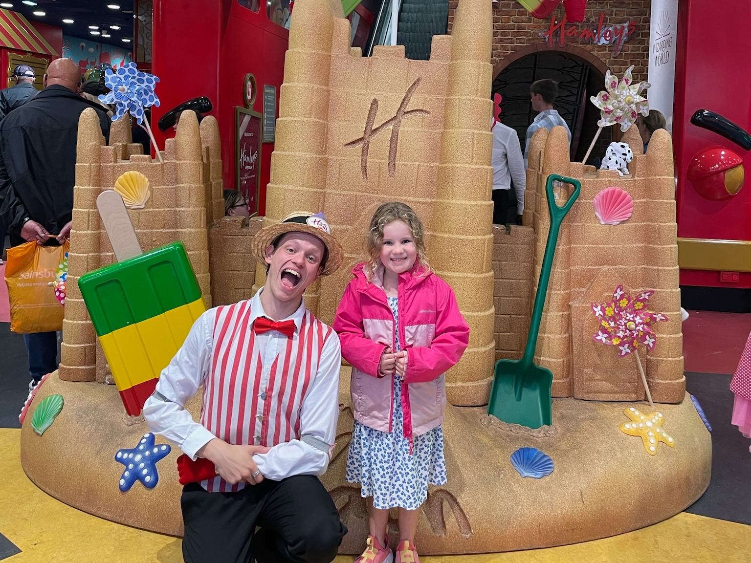 A child and an employee in front of a large sandcastle display inside a Hamleys toy store, surrounded by various toys and decorations.