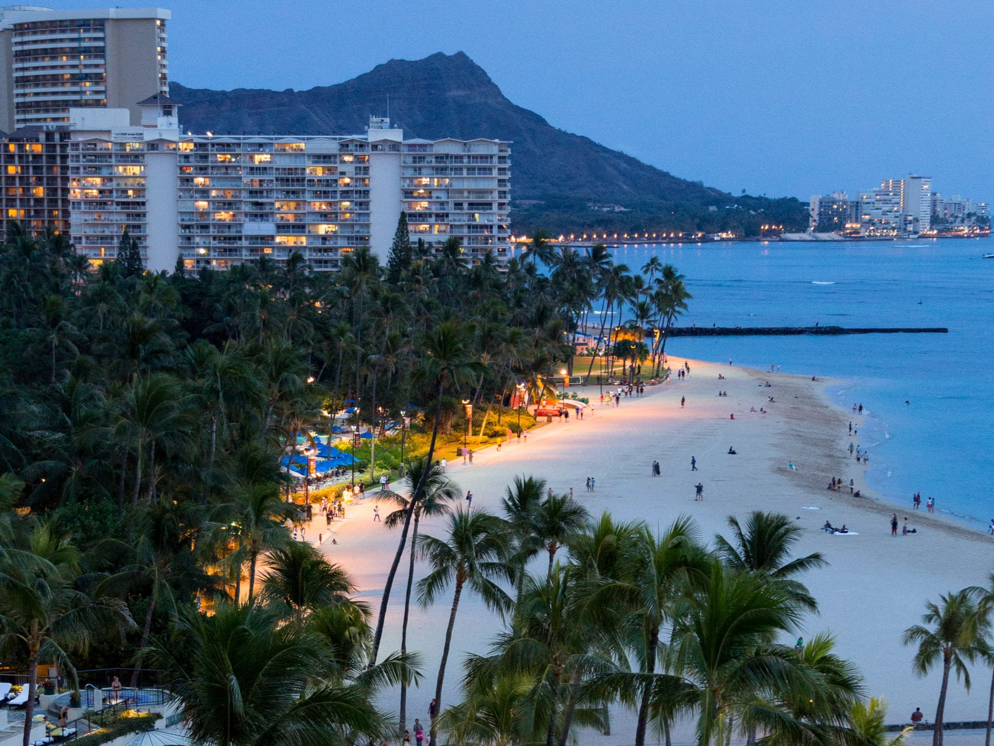 A scenic beachfront view at dusk with lit buildings, palm trees and a mountain in the background.