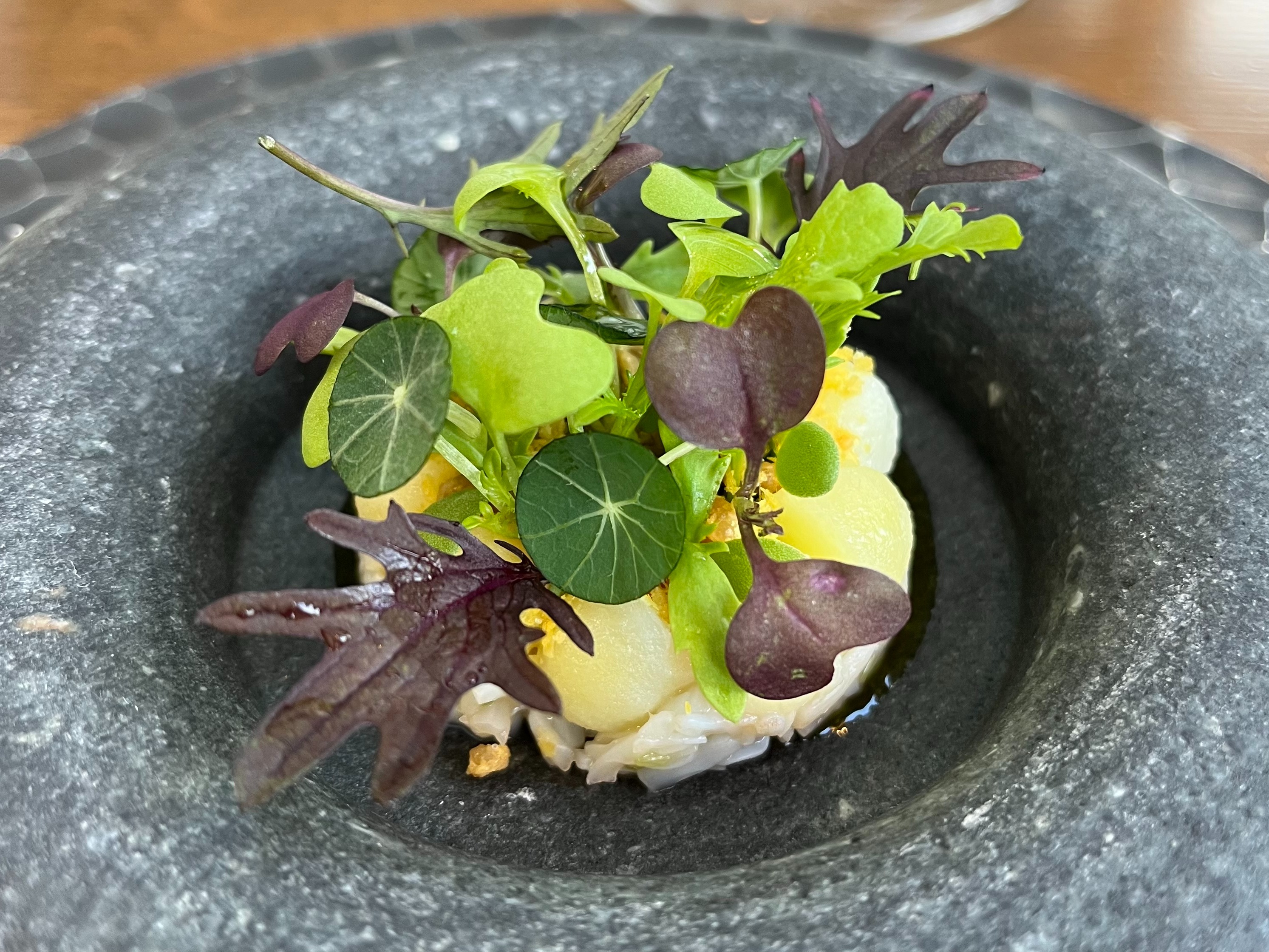 The image shows a gourmet dish with a variety of green and purple leaves atop a creamy base, served in a black bowl.