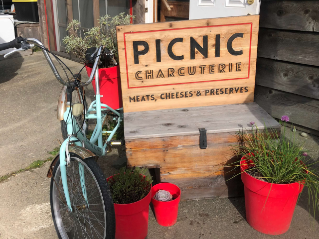 A light blue bicycle is parked next to a wooden bench with a sign advertising picnic charcuterie, surrounded by red potted plants.
