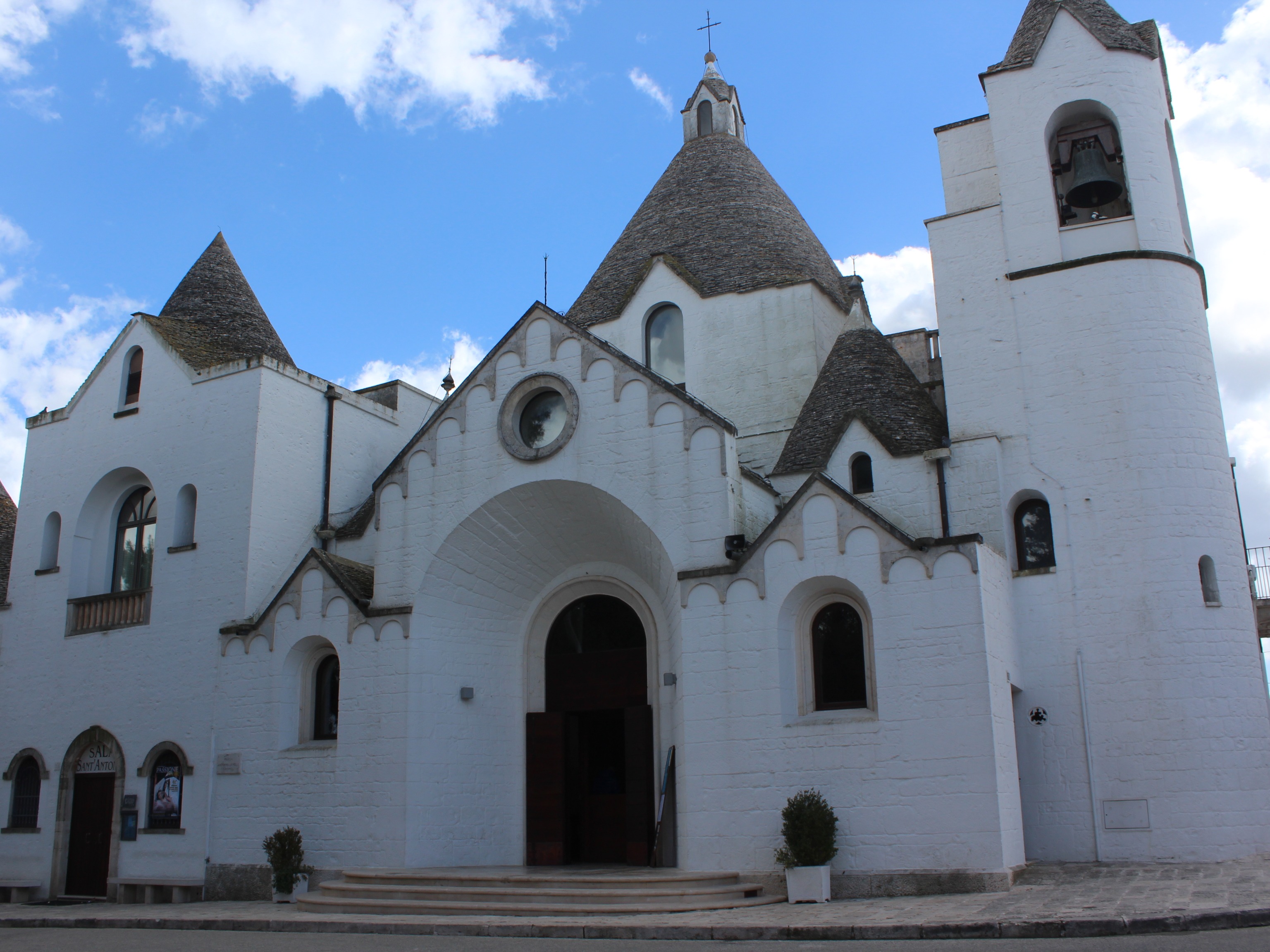 A white trullo church with a conical roof and bell tower under a blue sky with clouds.
