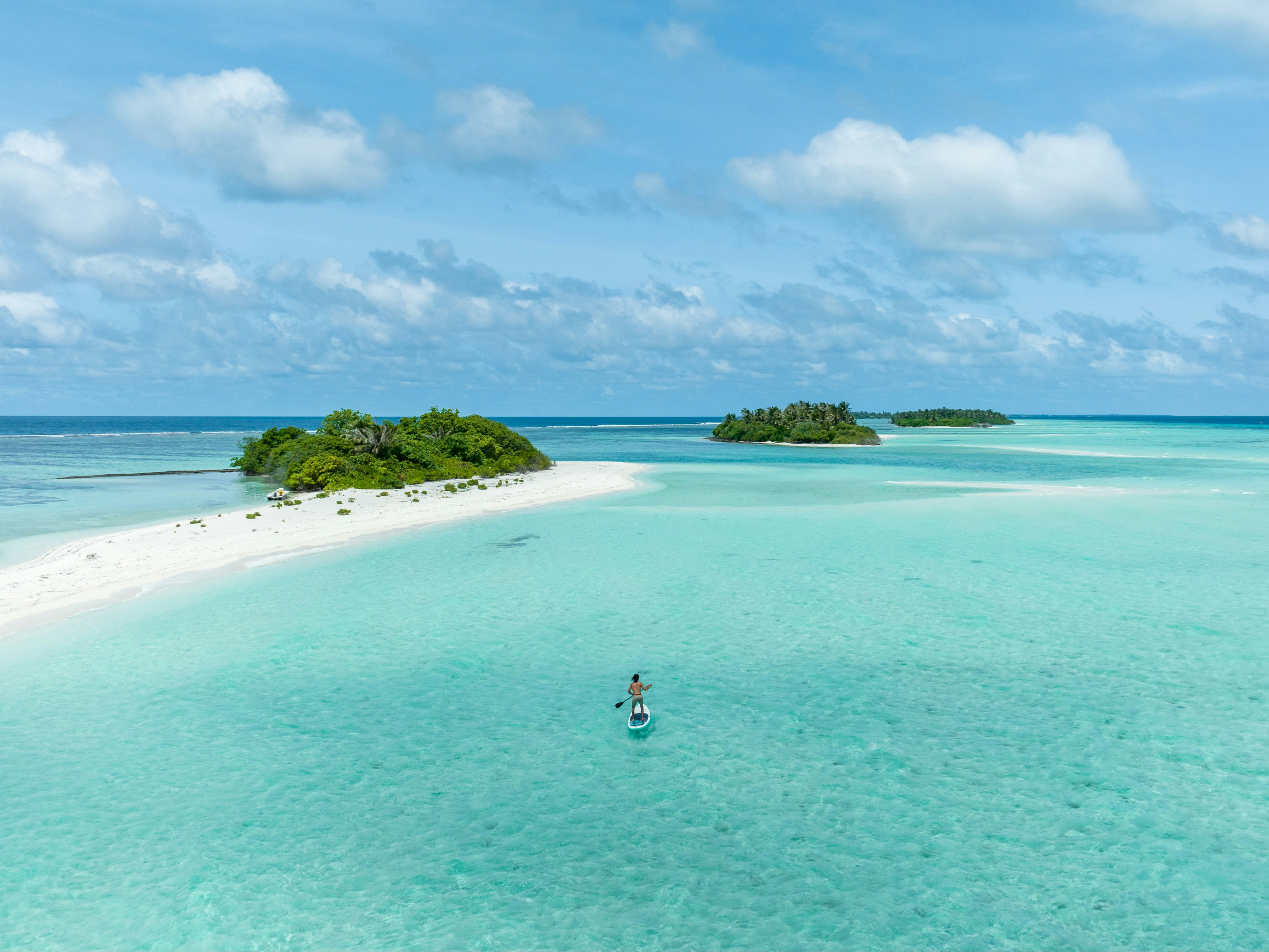 A person is paddleboarding in clear, shallow waters near a sandy beach with two small islands in the background.