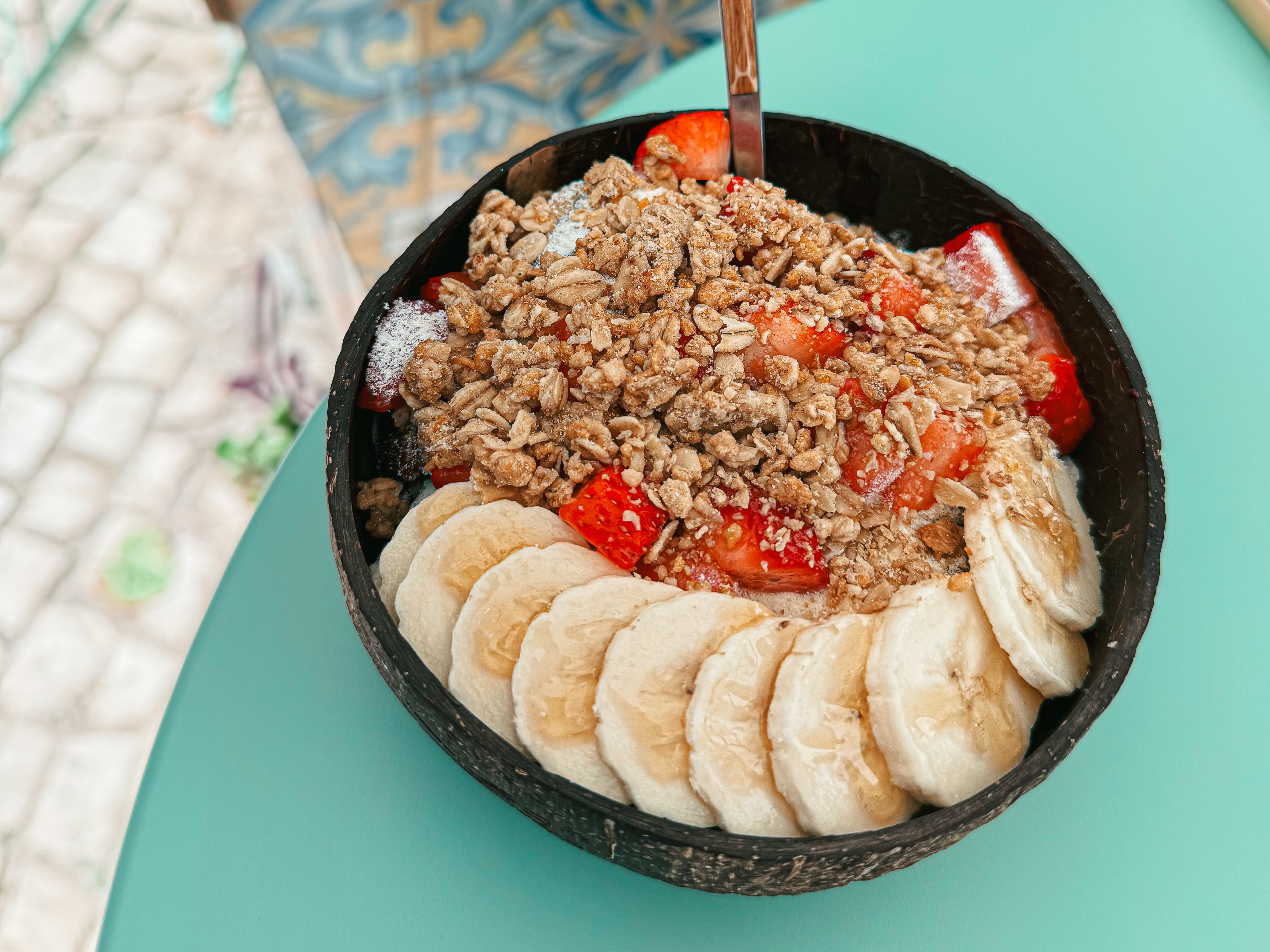 An aerial view of black bowl with a mix of granola and fruit on a light green table.