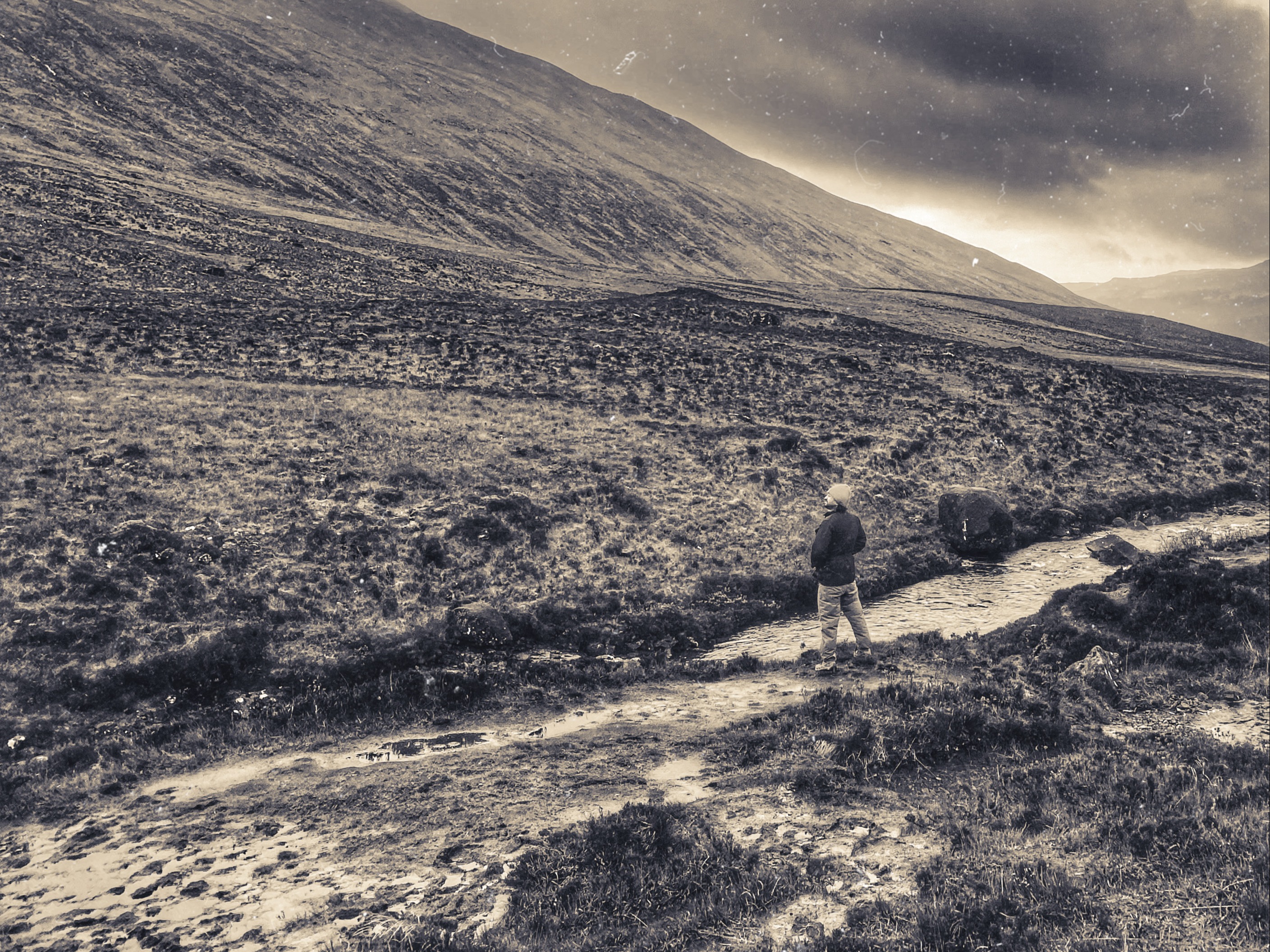 A person walks alone on a rugged path through hilly terrain under an overcast sky.