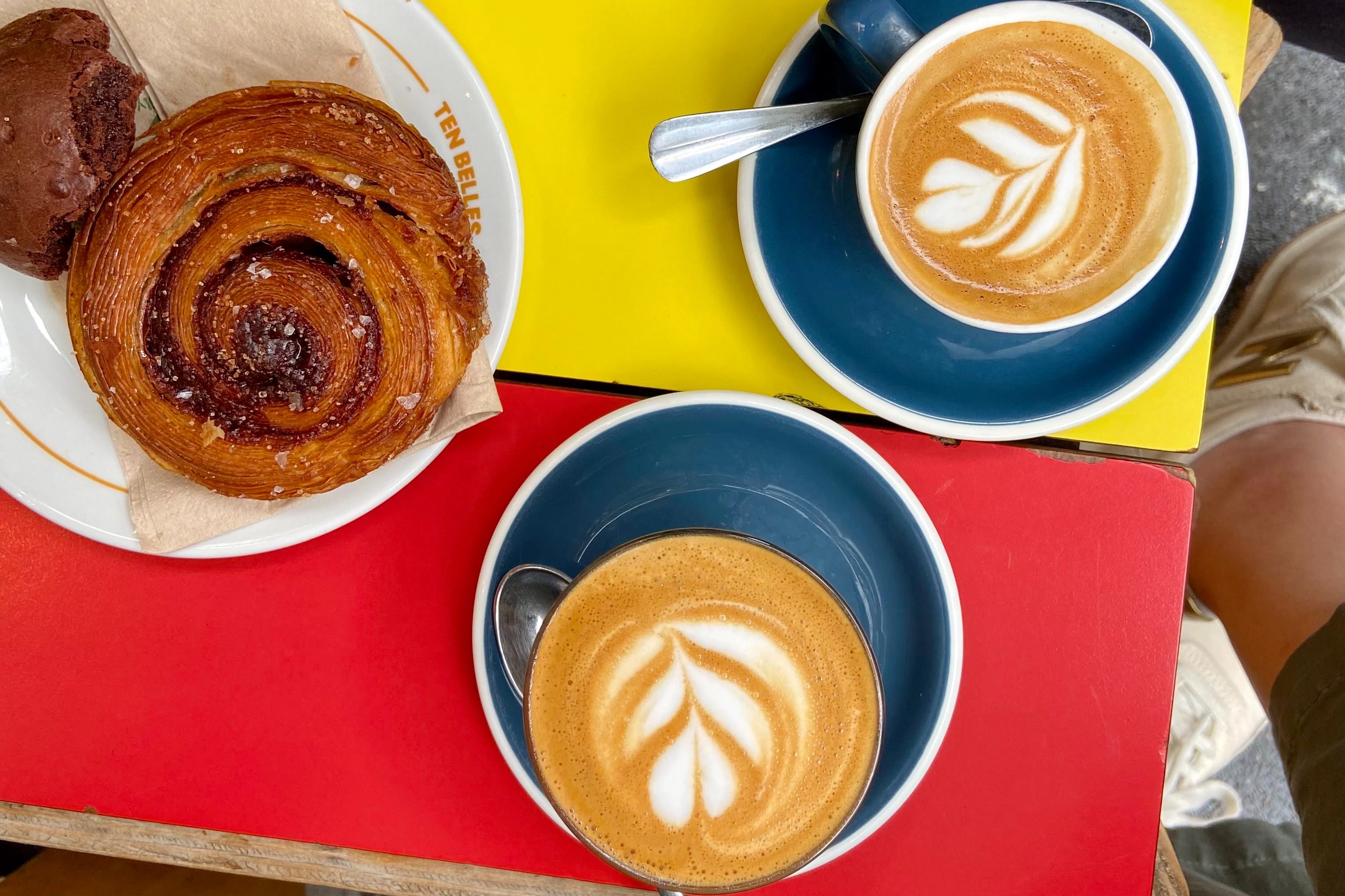 An aerial view of two coffees and a pastry on a yellow and red table at Ten Belles.