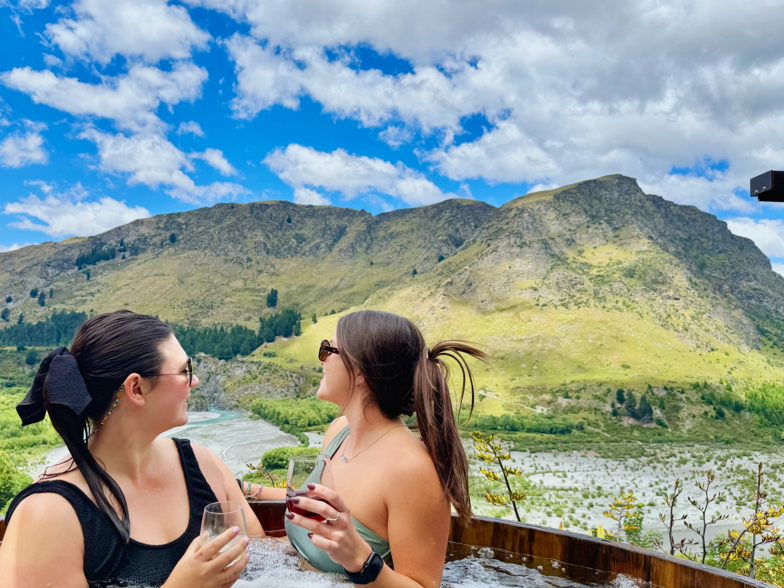Two women sitting in a hot tub, with a scenic backdrop of mountains and a river under a blue sky.