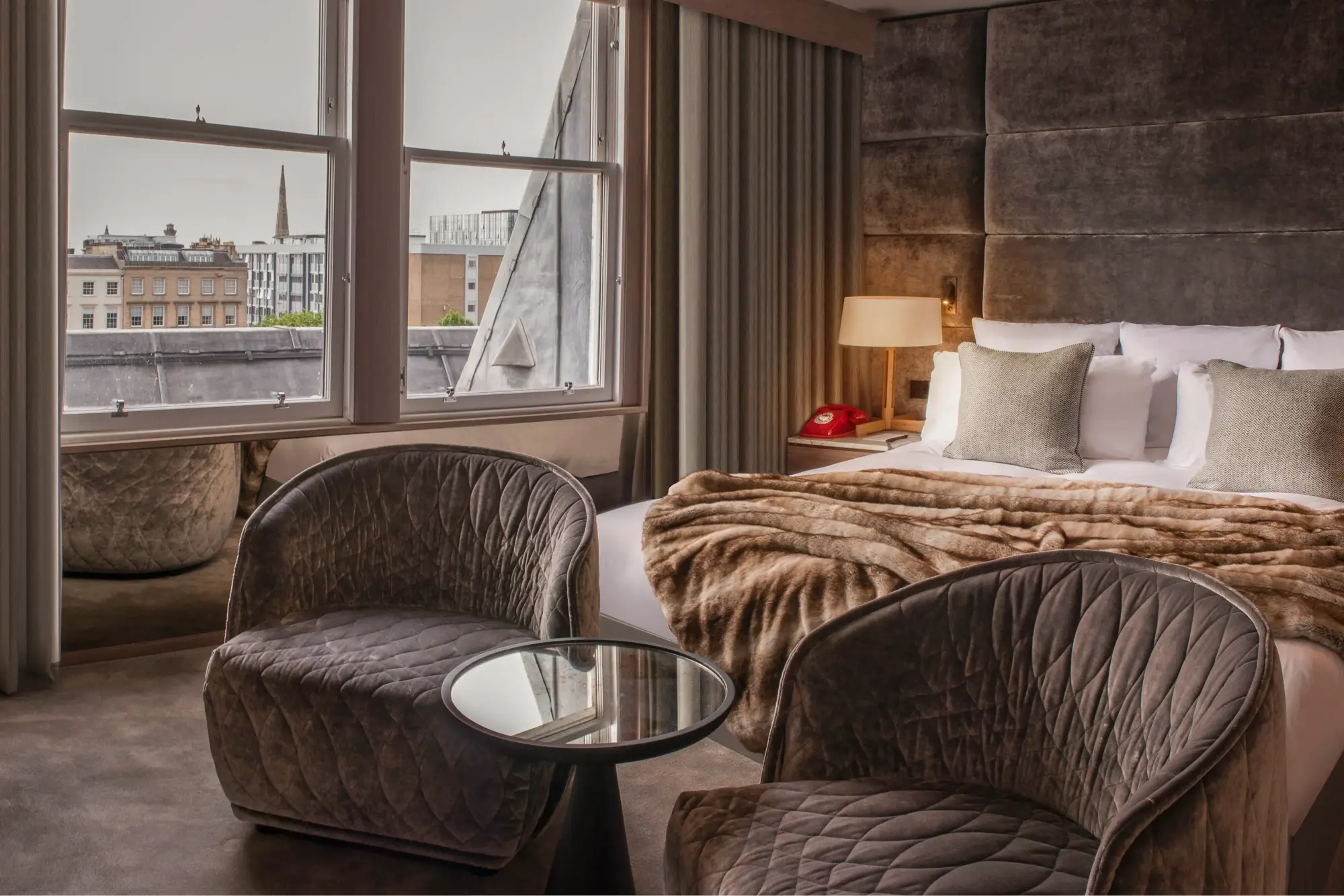 Stone walls, moody yet cozy furnishings fill a room at Kimpton Blythswood Squrare, with the city's historic skyline visible through large windows