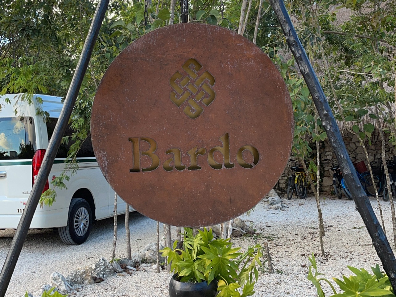 The image shows a circular rust-colored sign with the word “Bardo” cut out, hanging from a metal tripod structure over two potted plants, set against a backdrop of trees.