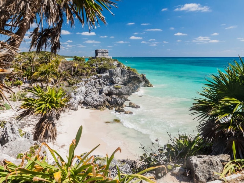 The image shows a scenic beach with turquoise waters, rocky cliffs, greenery, and an ancient stone structure atop the cliff under a clear blue sky.