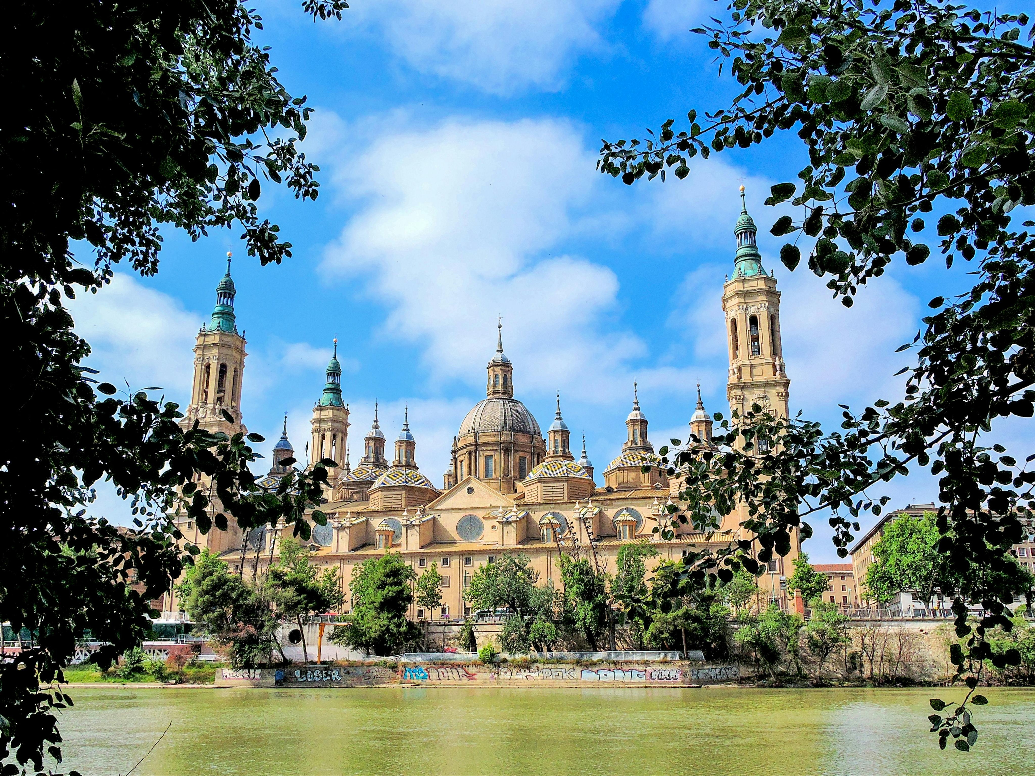 A grand cathedral with multiple domes and towers, viewed across a river and framed by foliage under a blue sky.