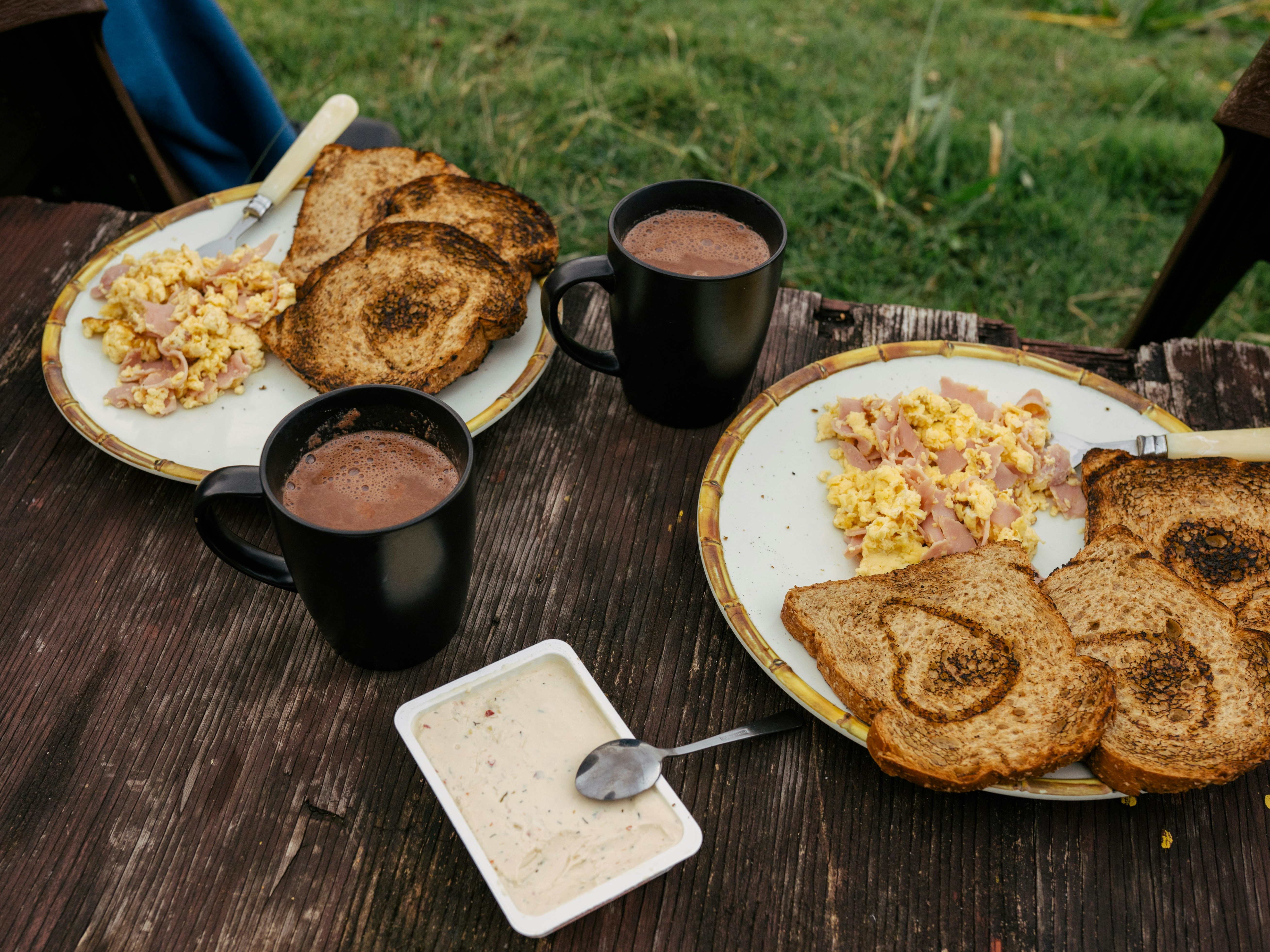 An outdoor meal with toasted bread, scrambled eggs and cups of hot beverage on a wooden table.