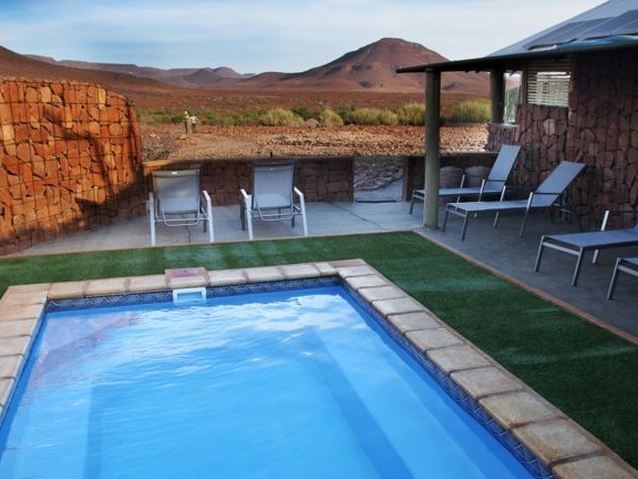 An outdoor swimming pool with a stone wall and chairs, set against a desert landscape.