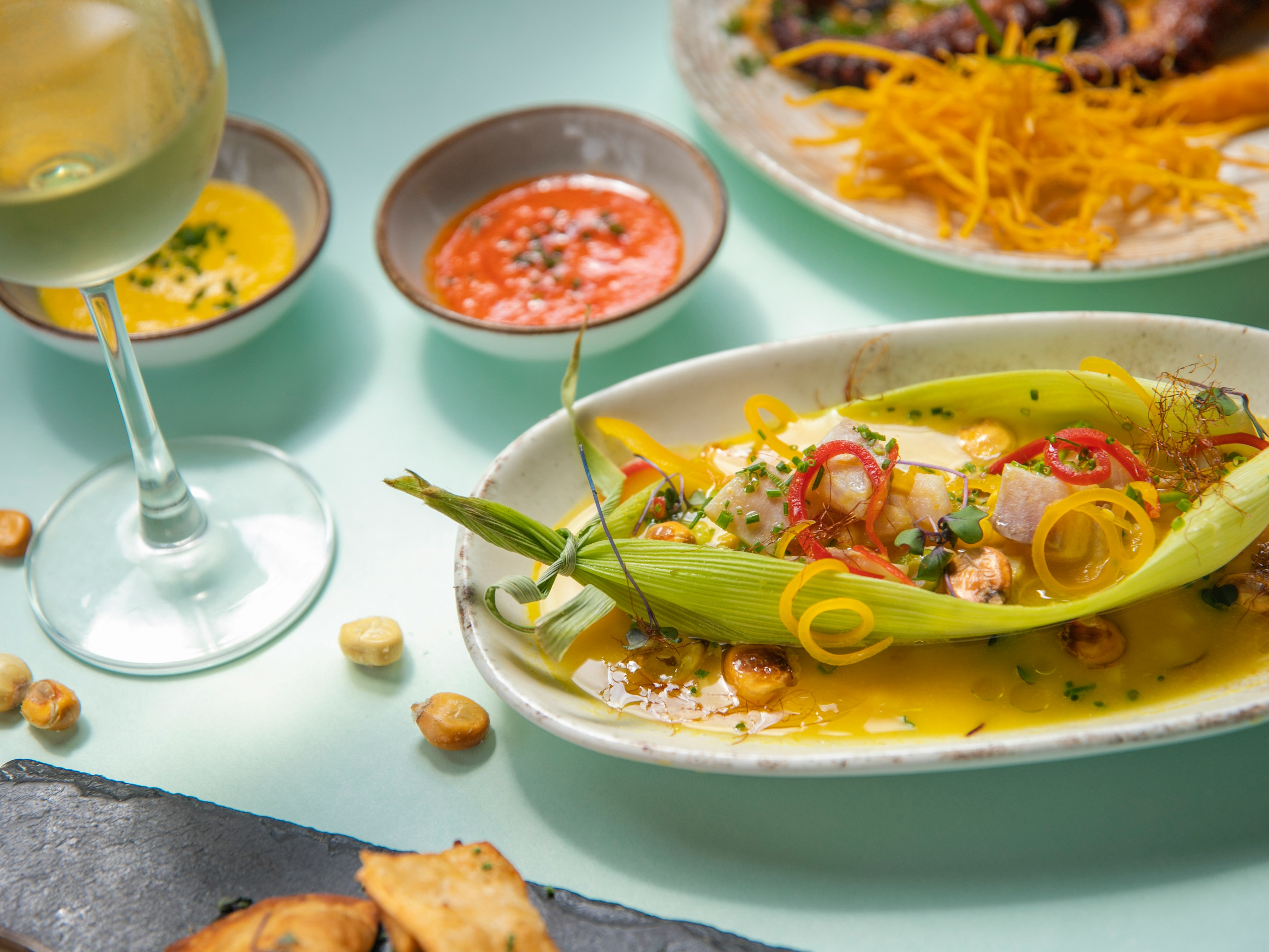 A plated dish with various vegetables and sauces, accompanied by a glass of wine and additional condiments on a light blue table.