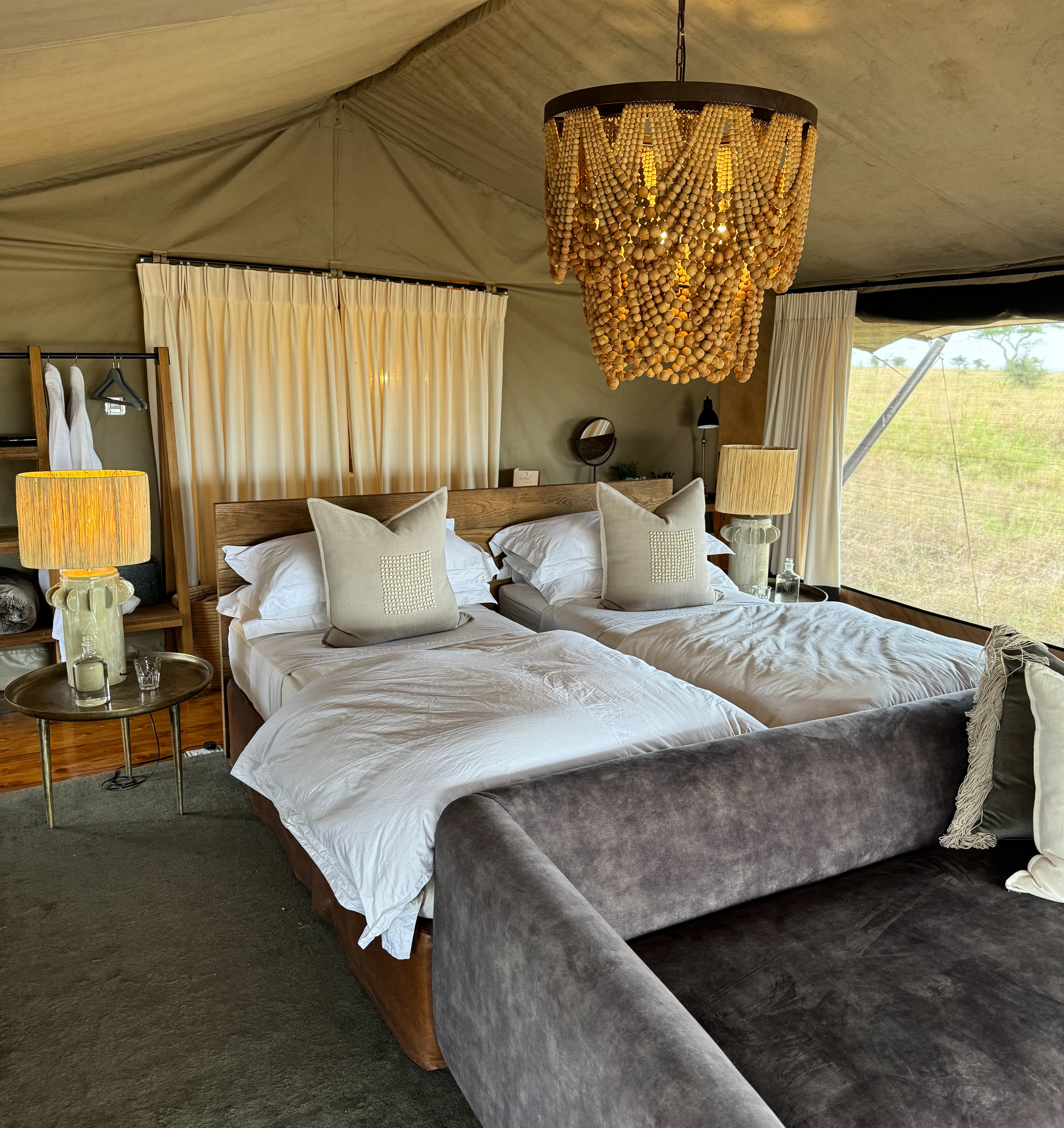 A view of a cozy hotel room with two twin beds, a grey couch and gold beaded chandelier in Tanzania.