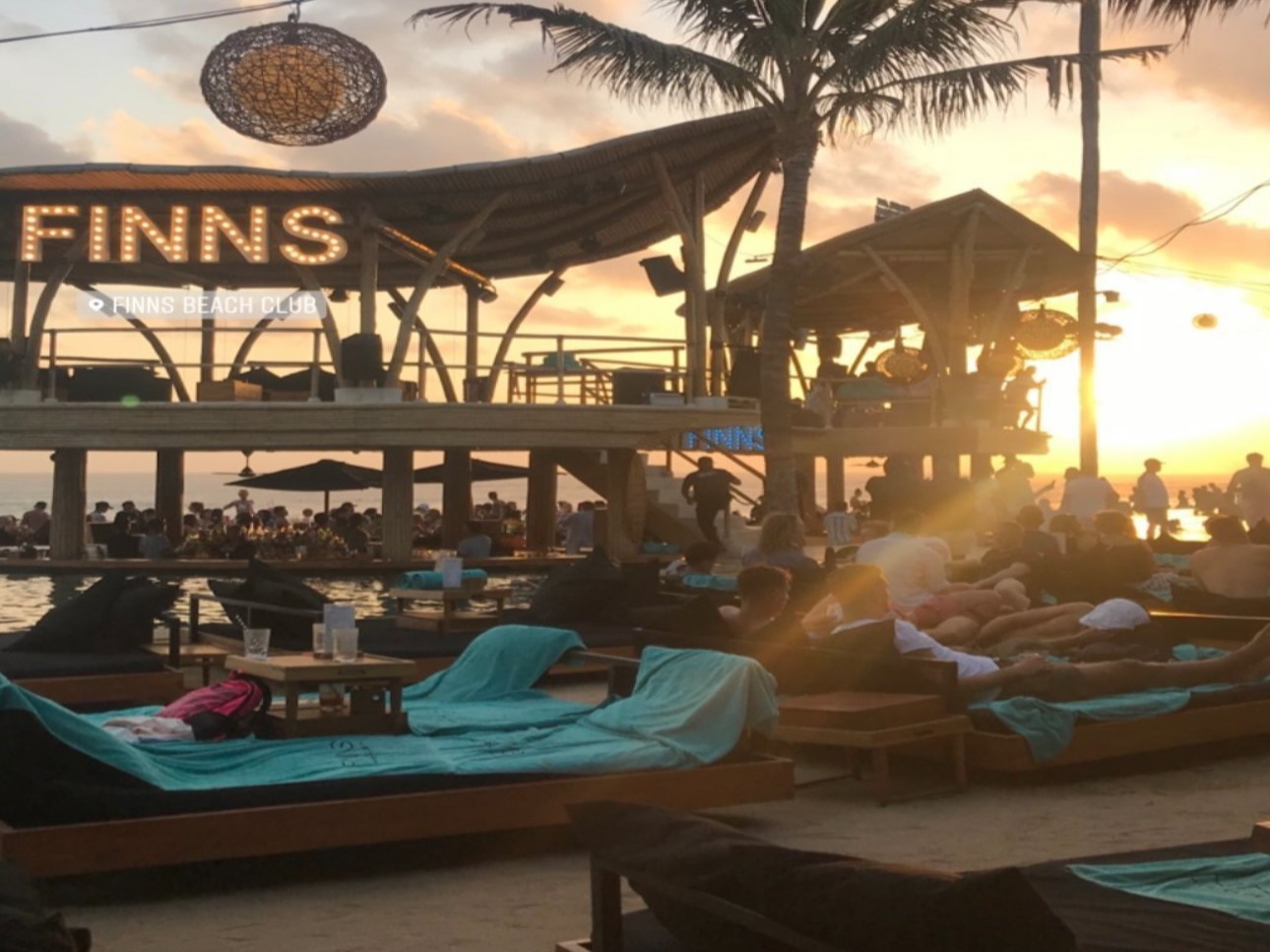 The image depicts a sunset view at a beach club named “FINNS” with people lounging and a silhouette of palm trees against the sky.