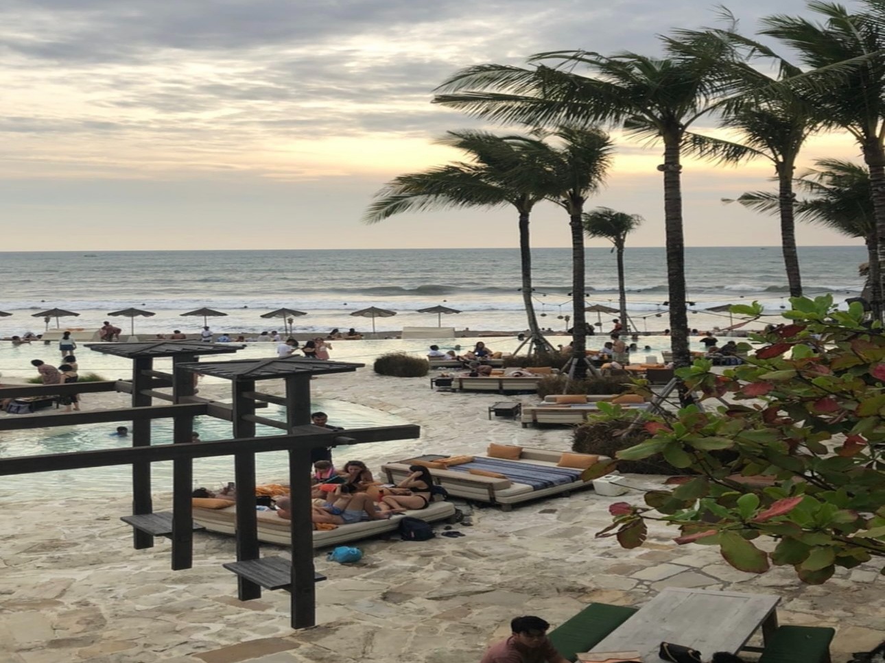 The image shows a beachside resort with people lounging and swimming, framed by palm trees under a cloudy sky.