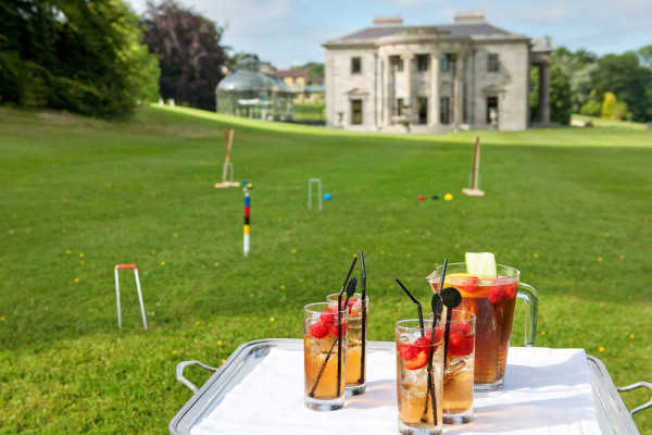 A jug and filled glasses on an outside table with a cricket set on a lawn
