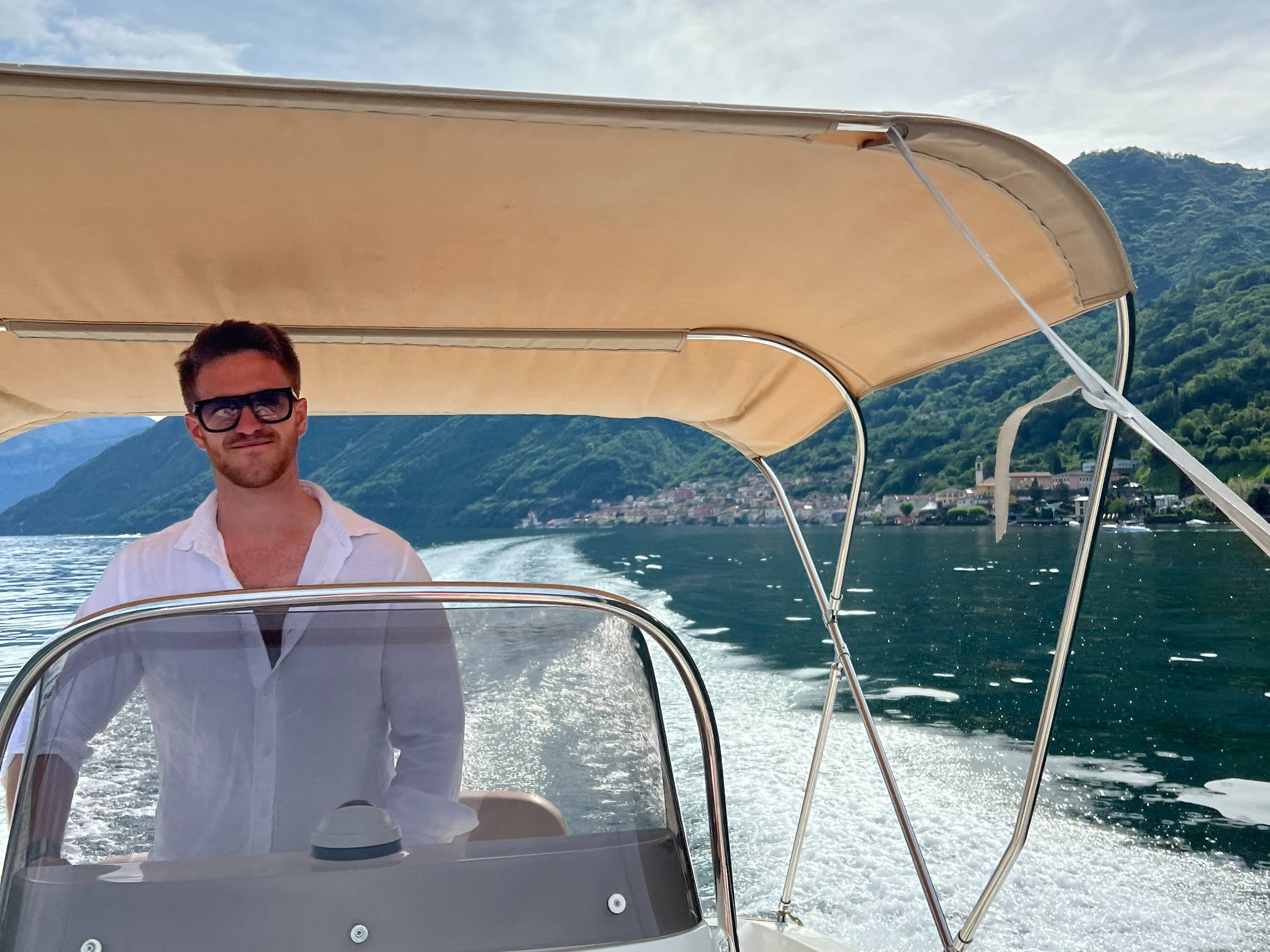 A person stands on a boat with a canopy, surrounded by water and mountains in the background.