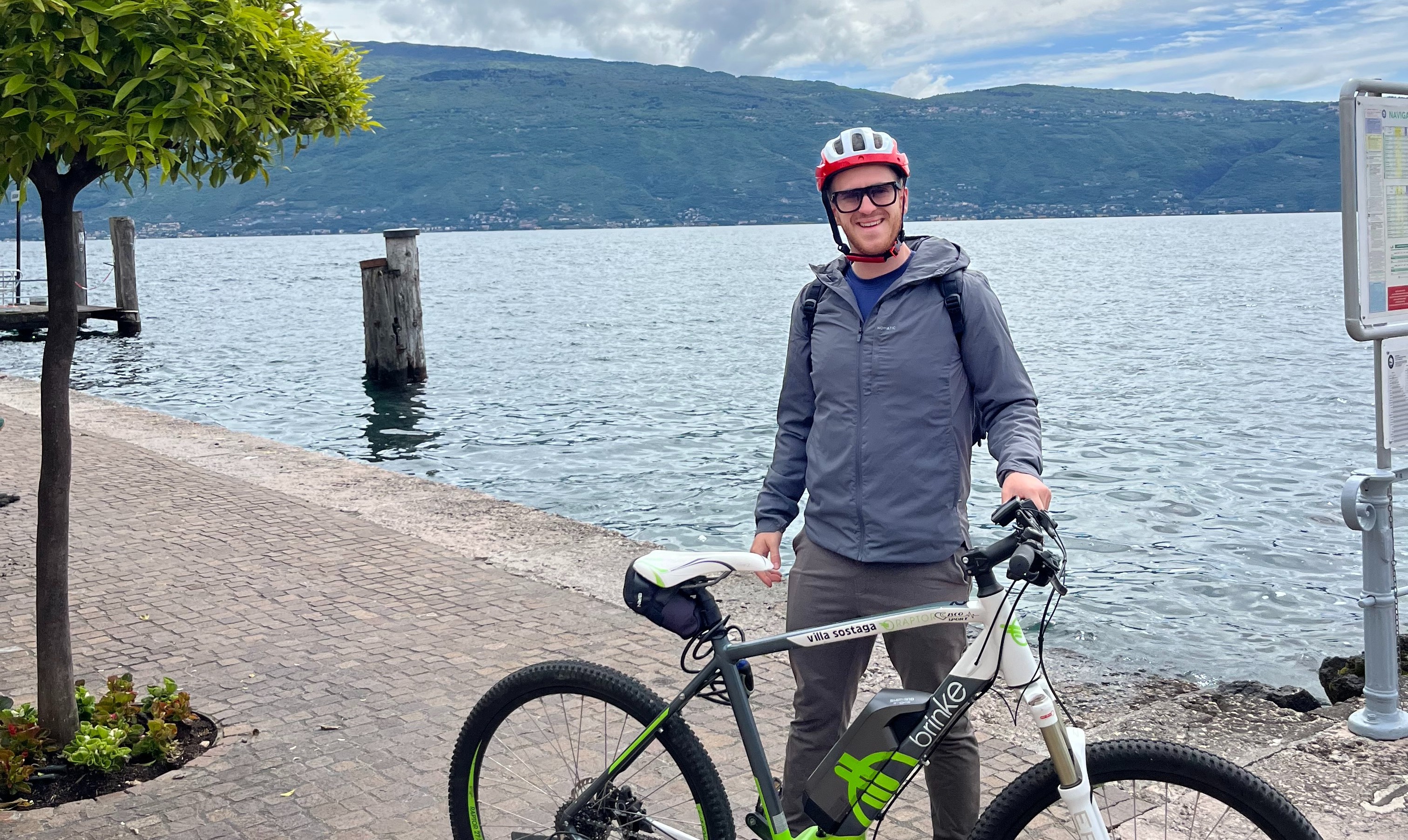 A person stands next to a bicycle by a lakeside with mountains in the background, under a partly cloudy sky.