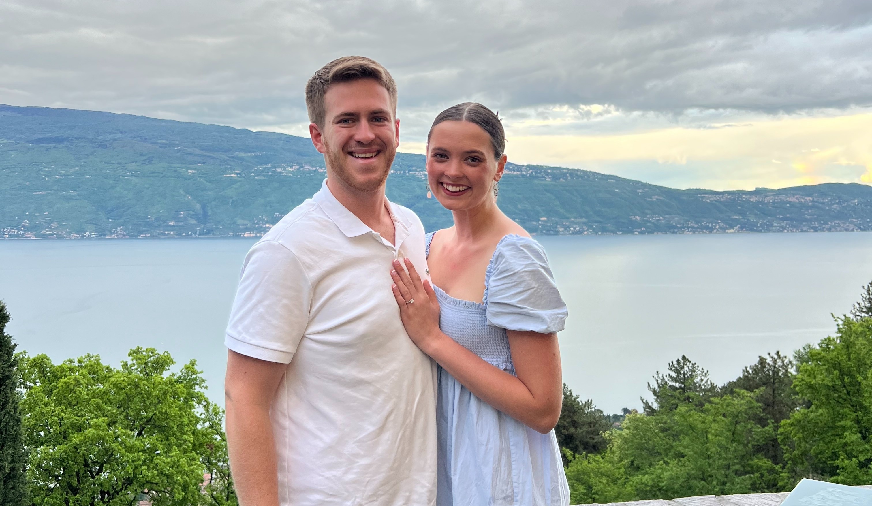 Two people pose for a photo with a scenic backdrop of hills and a cloudy sky.