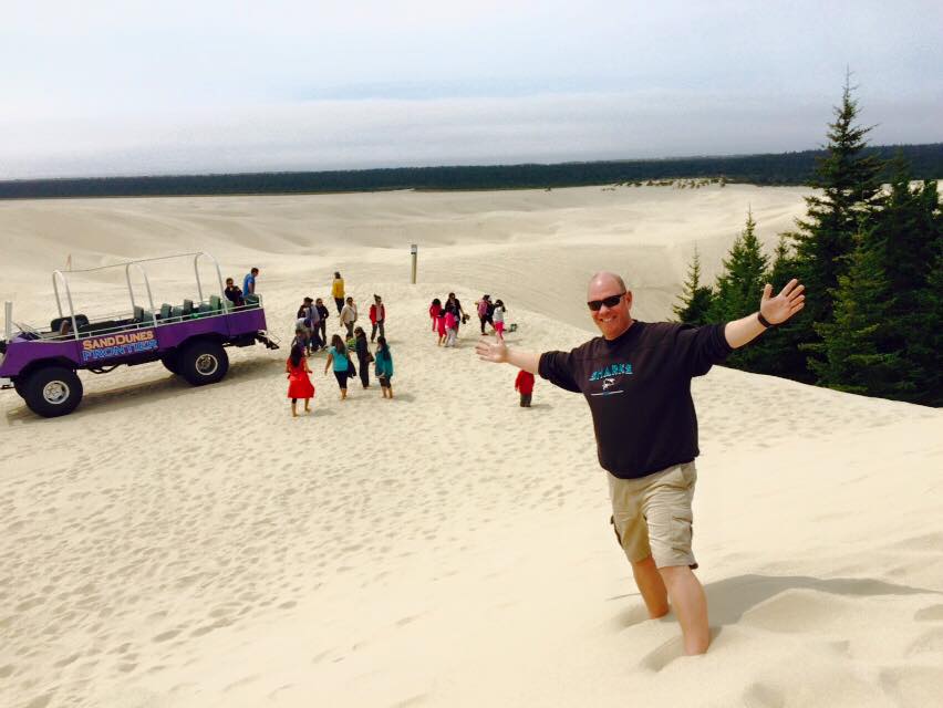 Travel advisor Dave standing on san dunes with a purple truck and people behind.