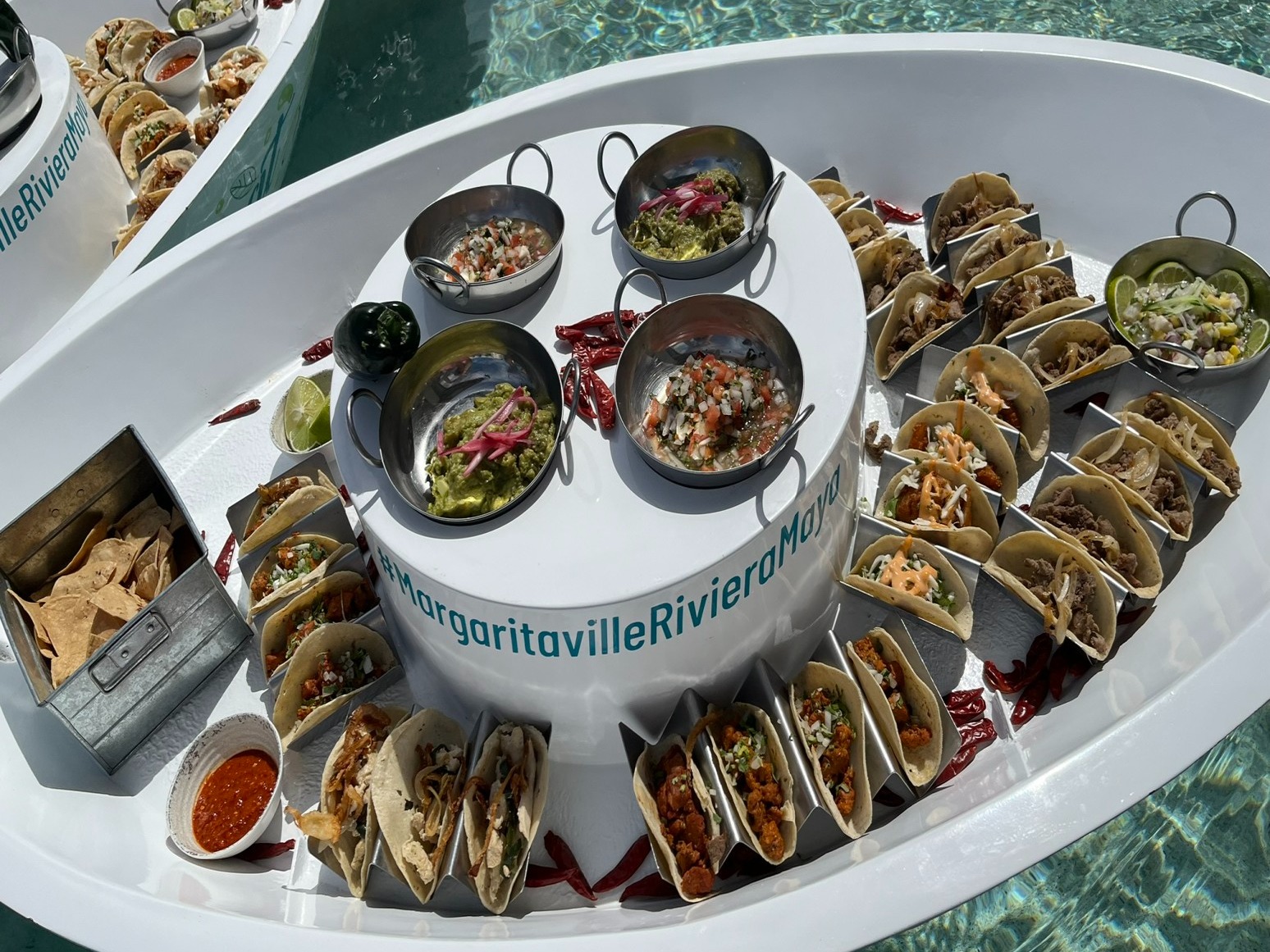 The image shows a floating circular white table with various Mexican dishes, including tacos and dips, in a pool with the text “Margaritaville Riviera Maya” on it.