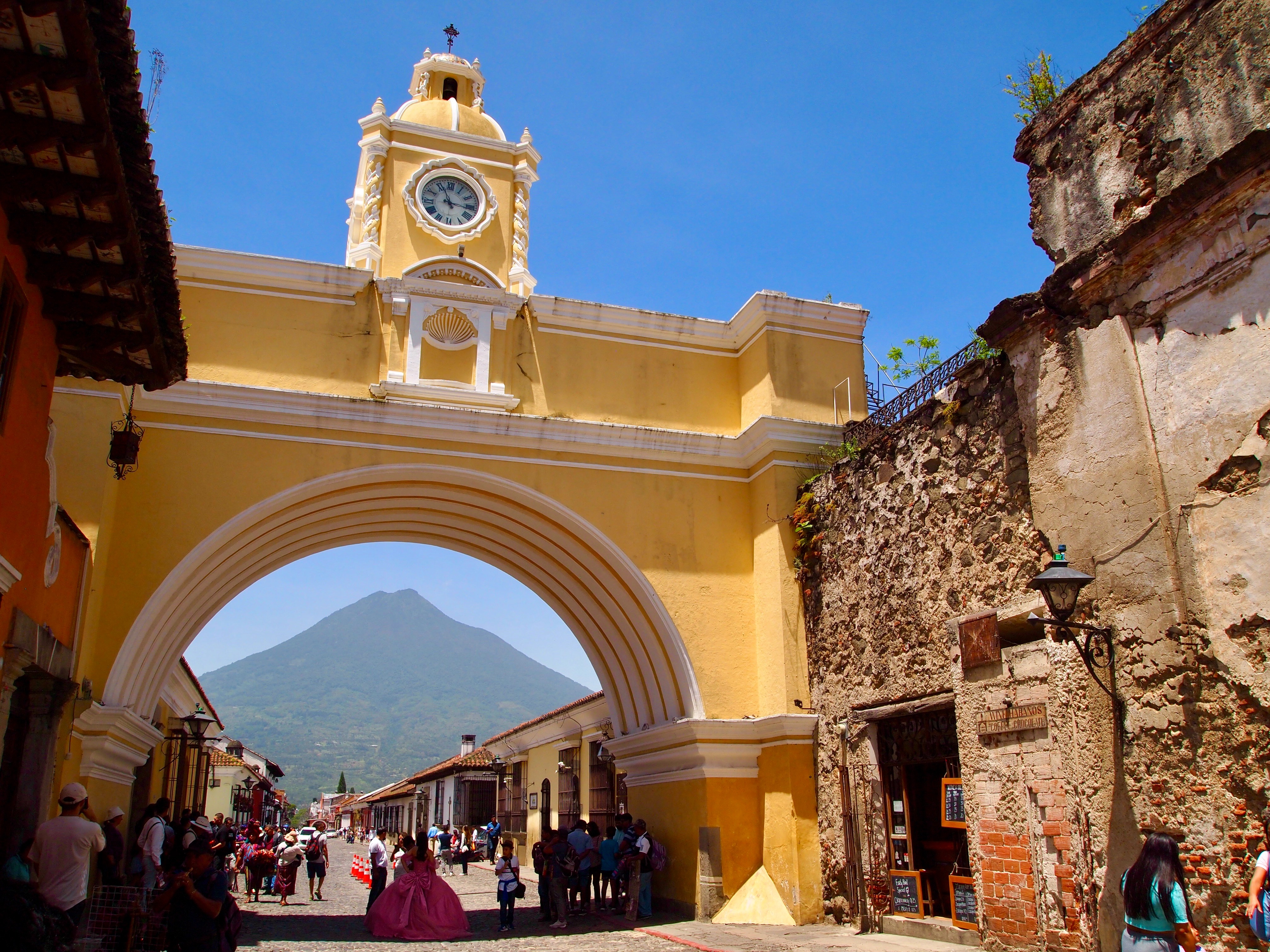 The image presents a lively street scene with people strolling under a prominent yellow archway, leading toward a distant volcano.