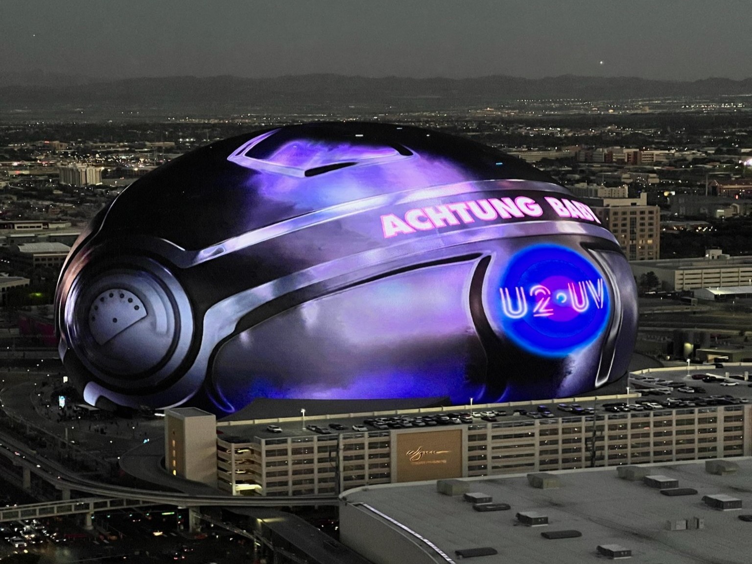 The image features a distinctive helmet-shaped building with “ACHTUNG BABY U2” lit up on its side during twilight in a cityscape.