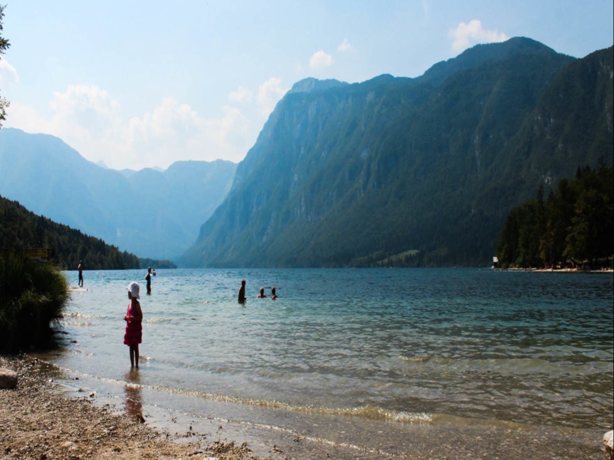 The image depicts a serene lakeside scene with individuals wading and mountains in the distance on a sunny day.