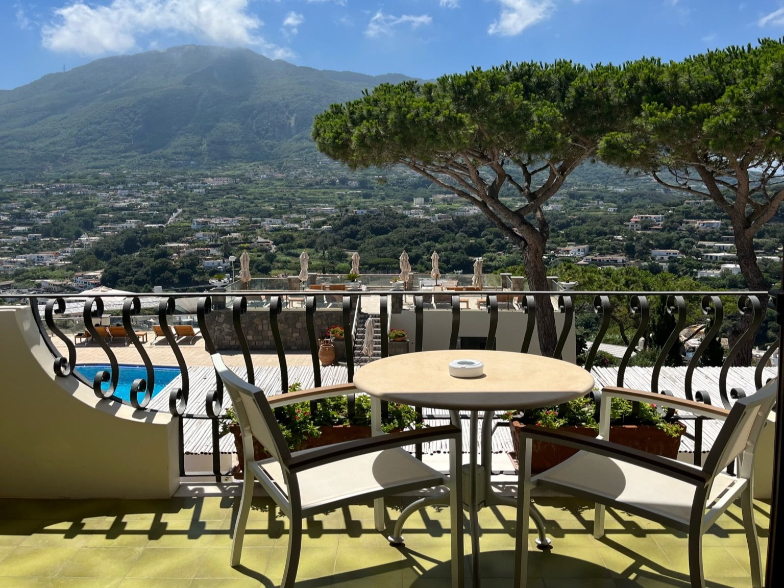 The image displays a scenic balcony view overlooking a mountainous landscape with lush greenery and a clear sky.
