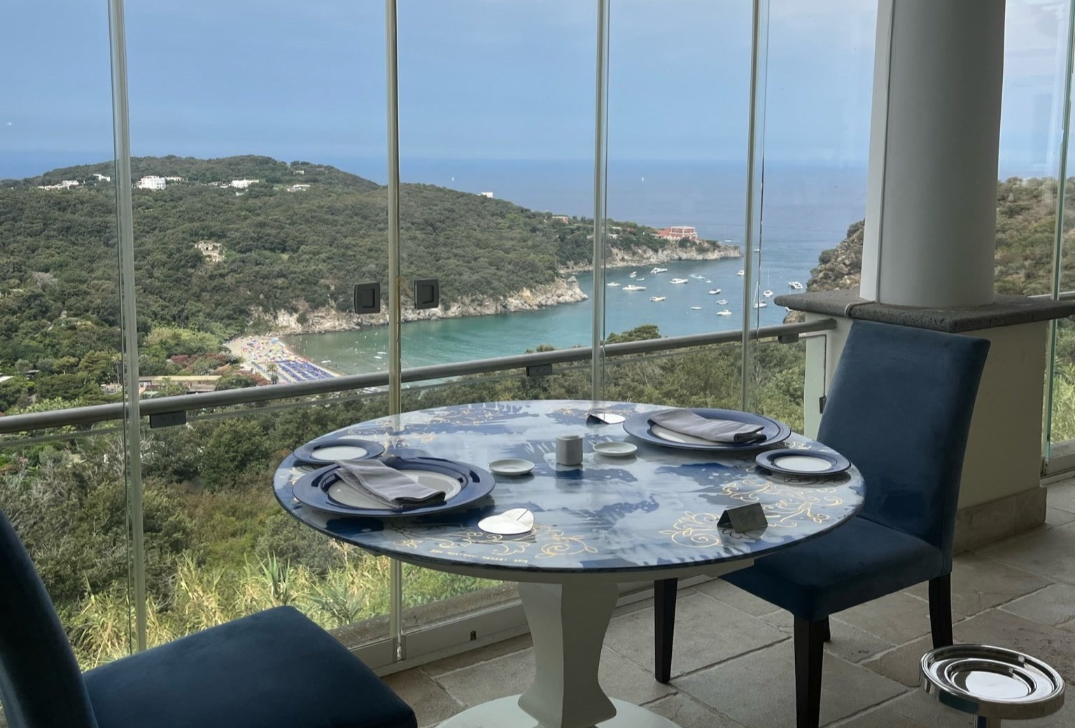 The image shows a dining table set for two, with a scenic view of a coastal landscape through floor-to-ceiling windows.