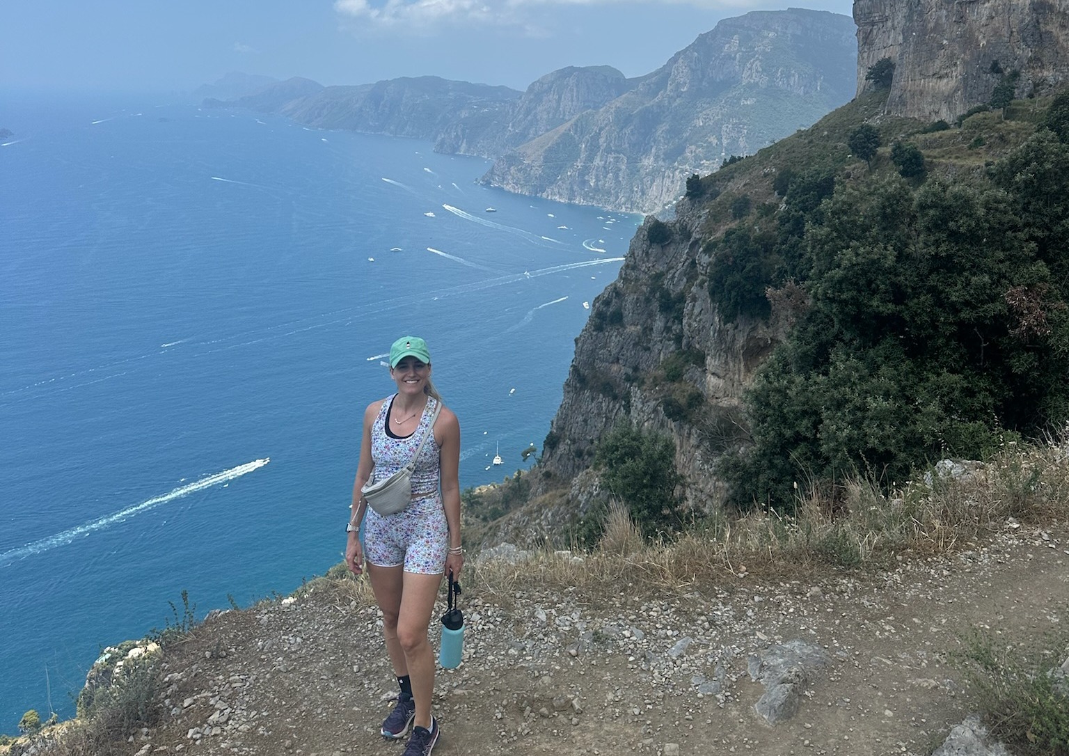 The image shows an individual walking on a coastal trail with steep cliffs and a vast ocean in the background.