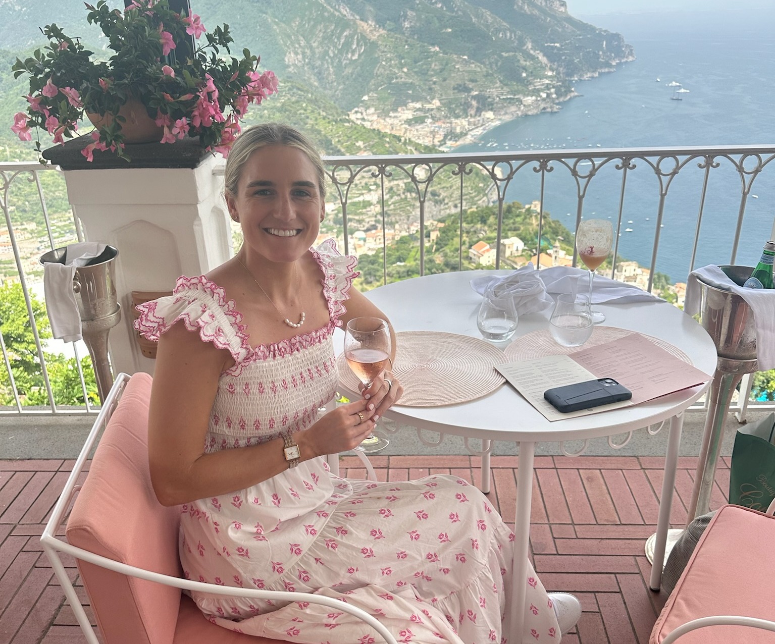 The image shows an individual sitting at a table with a glass in hand, overlooking a scenic coastal landscape from a balcony.