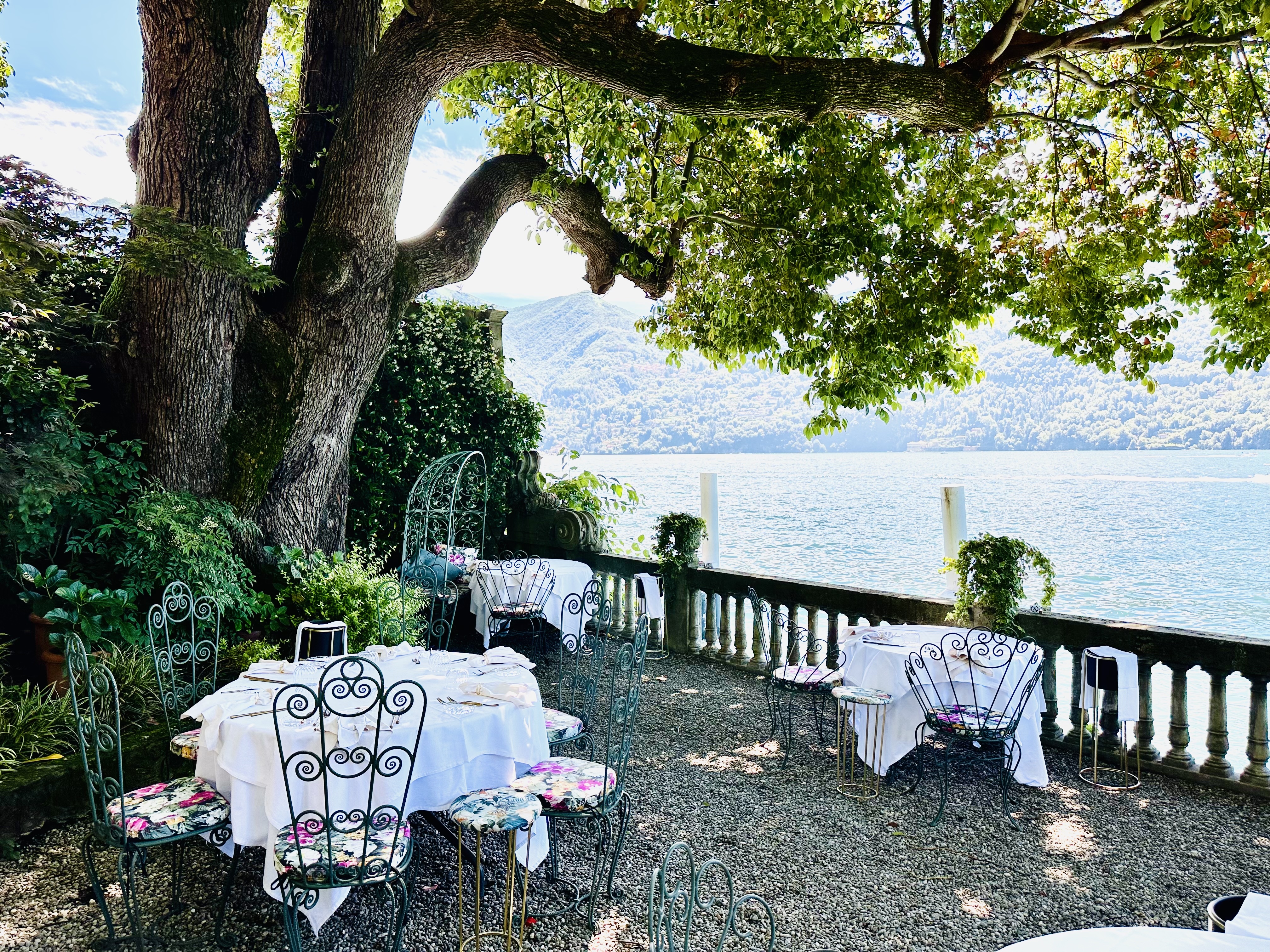 A view of an elegant restaurant terrace with three tables overlooking Lake Como, under a large tree.