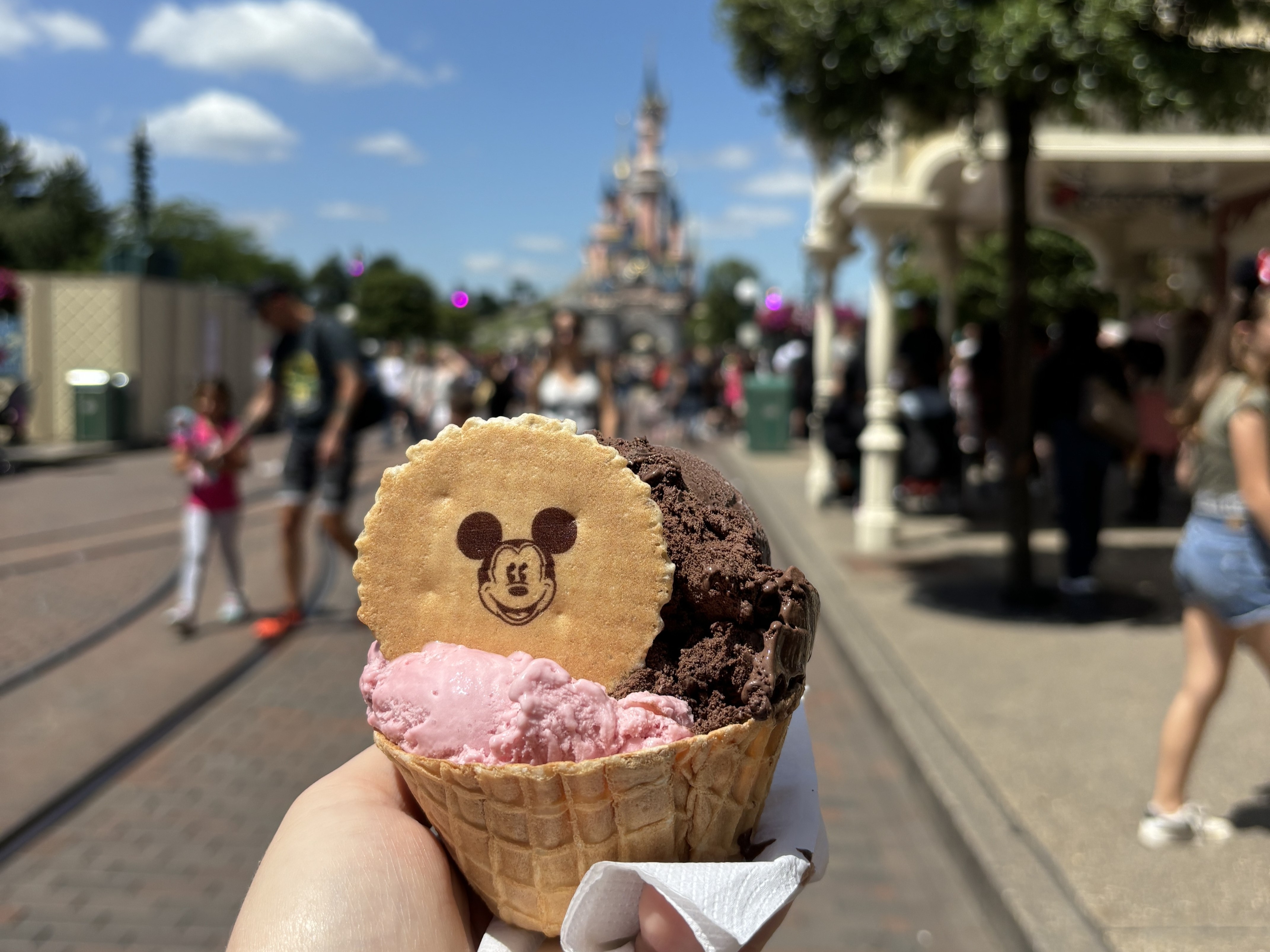 The image features a hand holding a waffle cone with two scoops of ice cream and a Mickey Mouse-shaped biscuit, against the backdrop of the Disneyland castle and visitors.