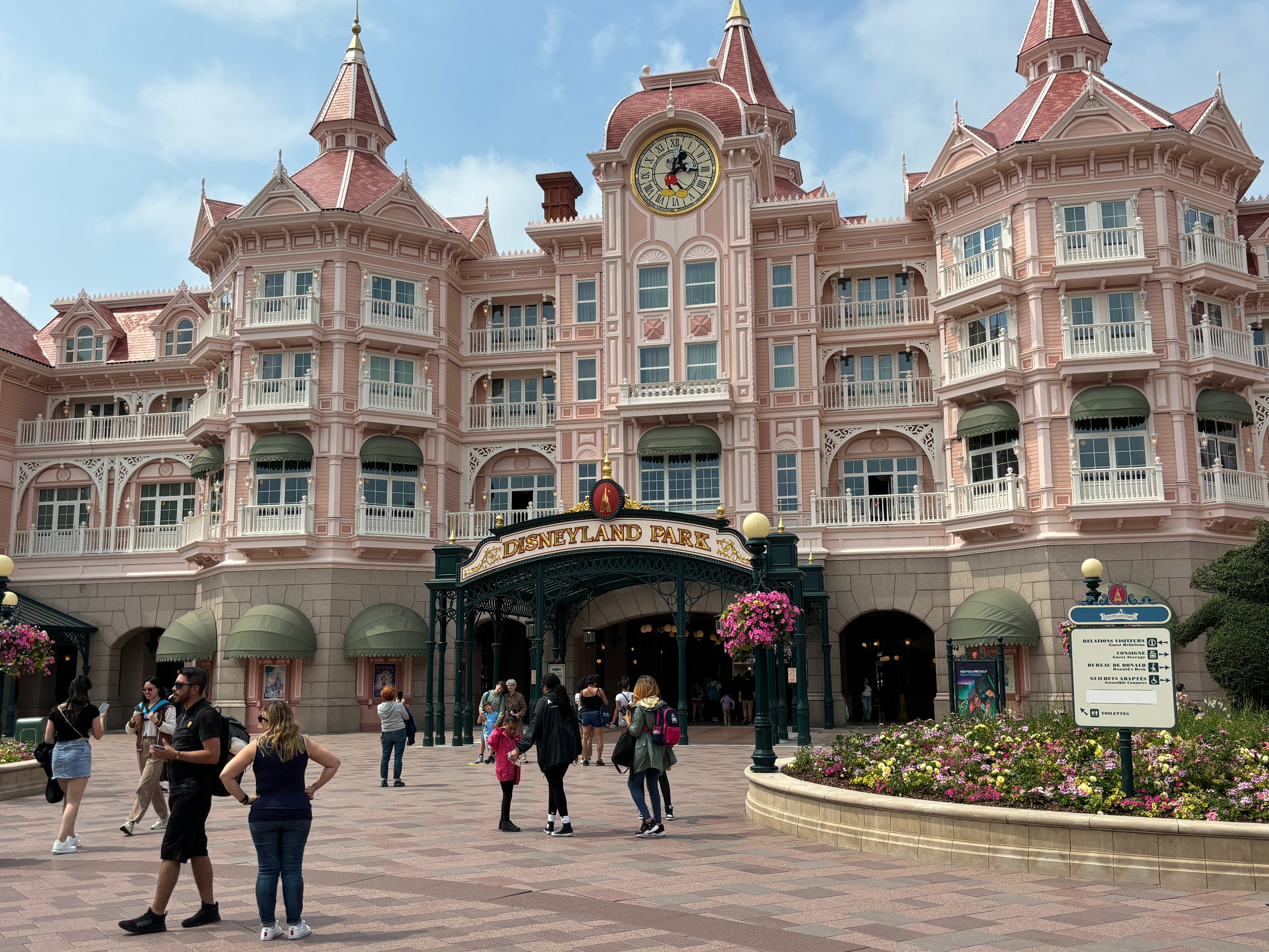 The image features a group of visitors in front of the pink-hued Disneyland Hotel with ornate Victorian architecture and a central clock tower under a blue sky.
