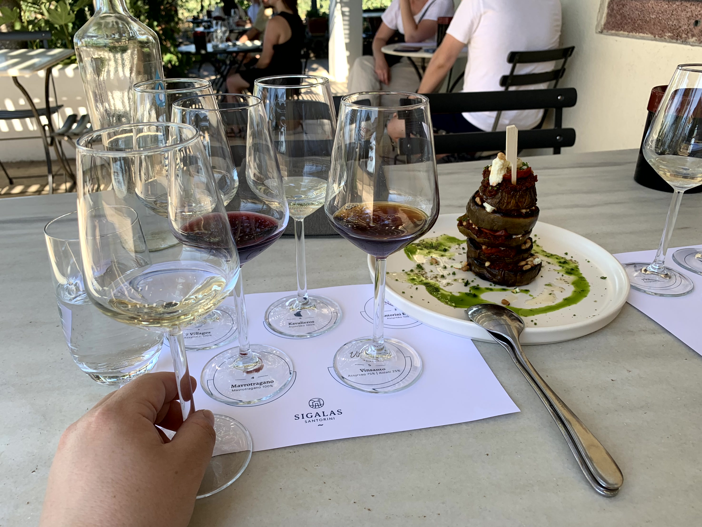 Multiple glasses for wine tasting arrayed on a patio table with dessert and silverware on a sunny day.