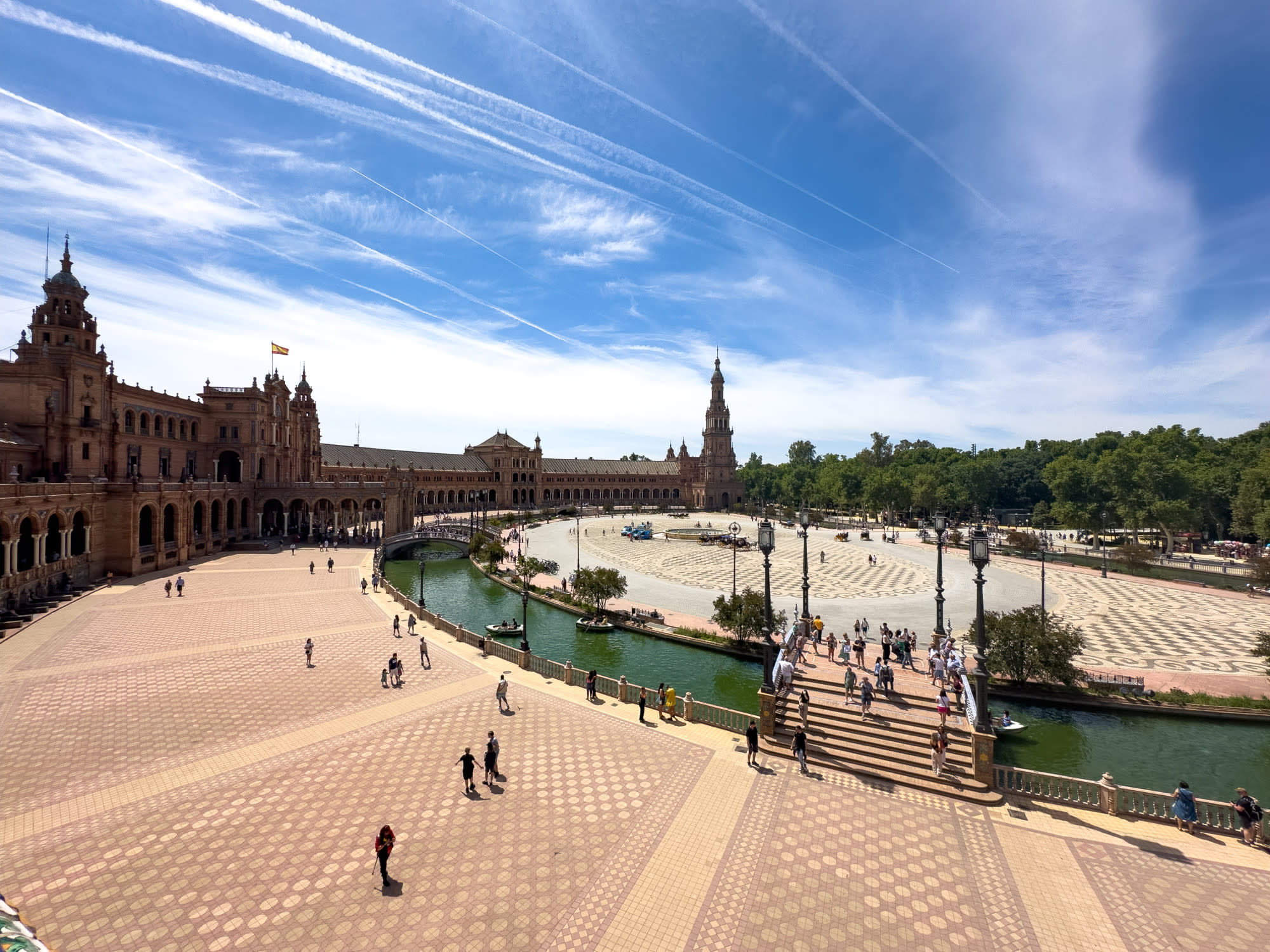 Bird's eye view of a Seville plaza with fountains and sparse greenery under a clear sky with wisps of clouds.
