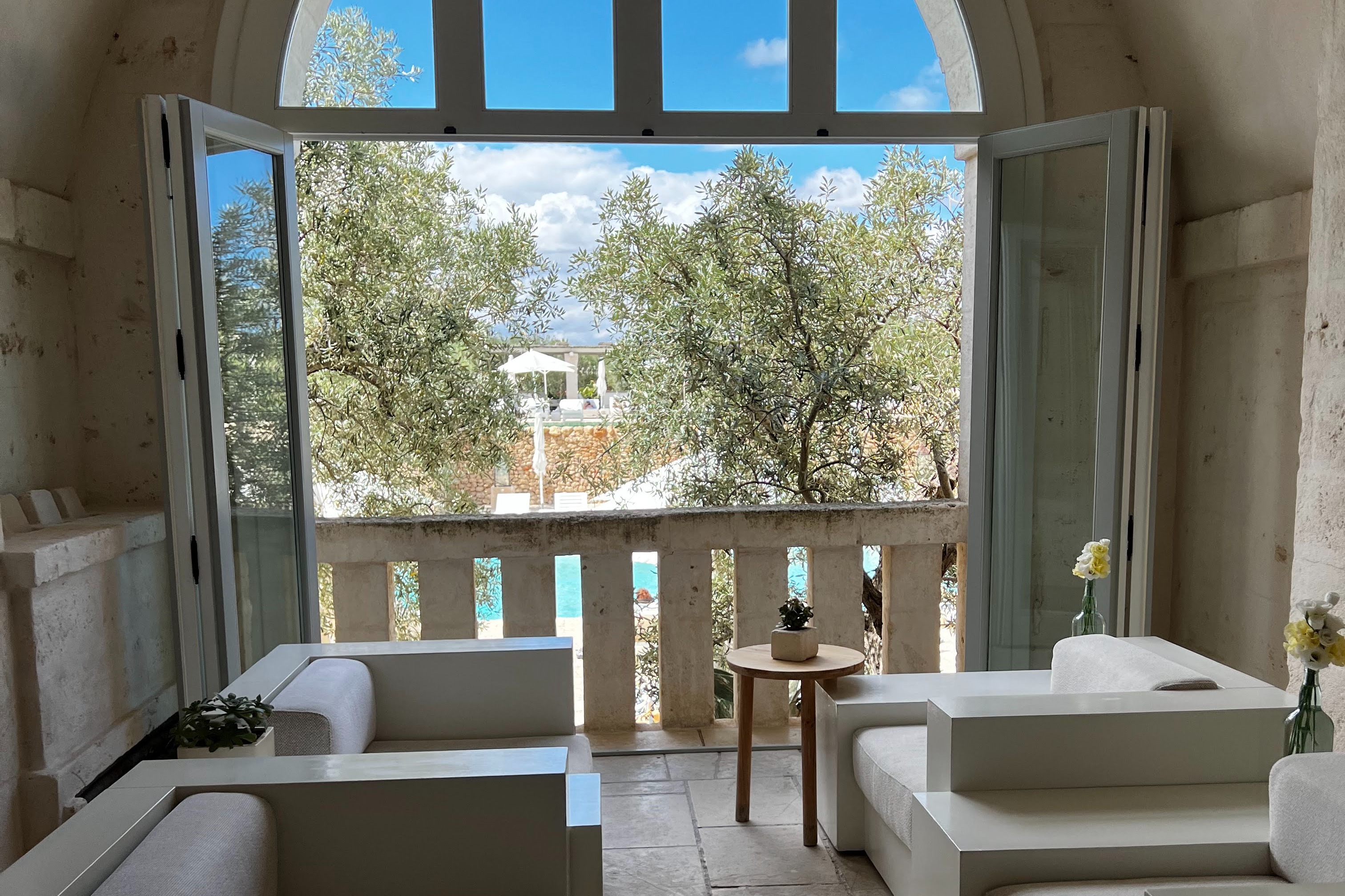 A view from a clean, white, minimal hotel room overlooking a lush pool area and ornate buildings in Borgo Egnazia.