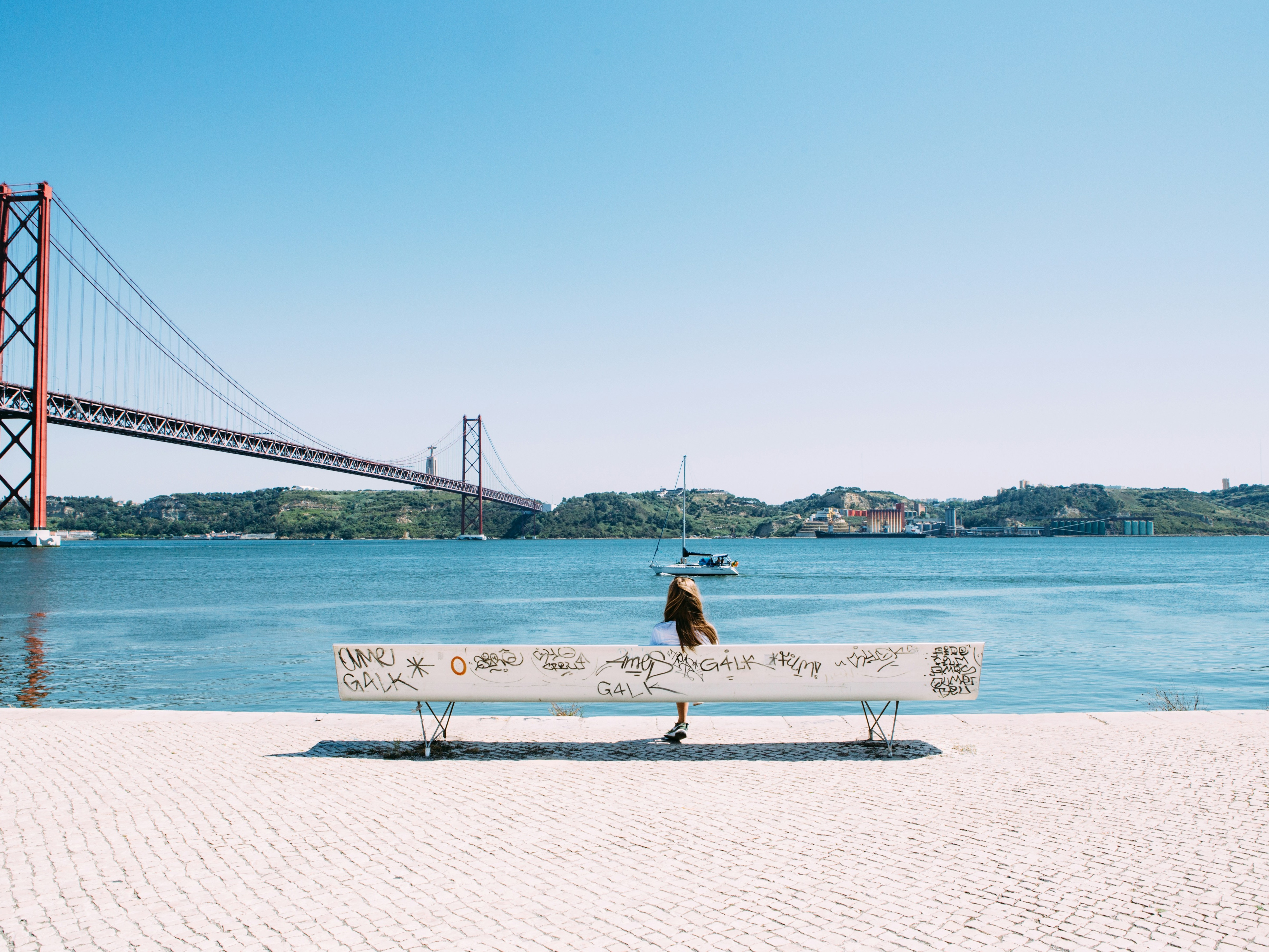 A person sitting on a bench adorned with graffiti, with a majestic suspension bridge that looks like the Golden Gate Bridge stretching across the water to low hills on the other side.