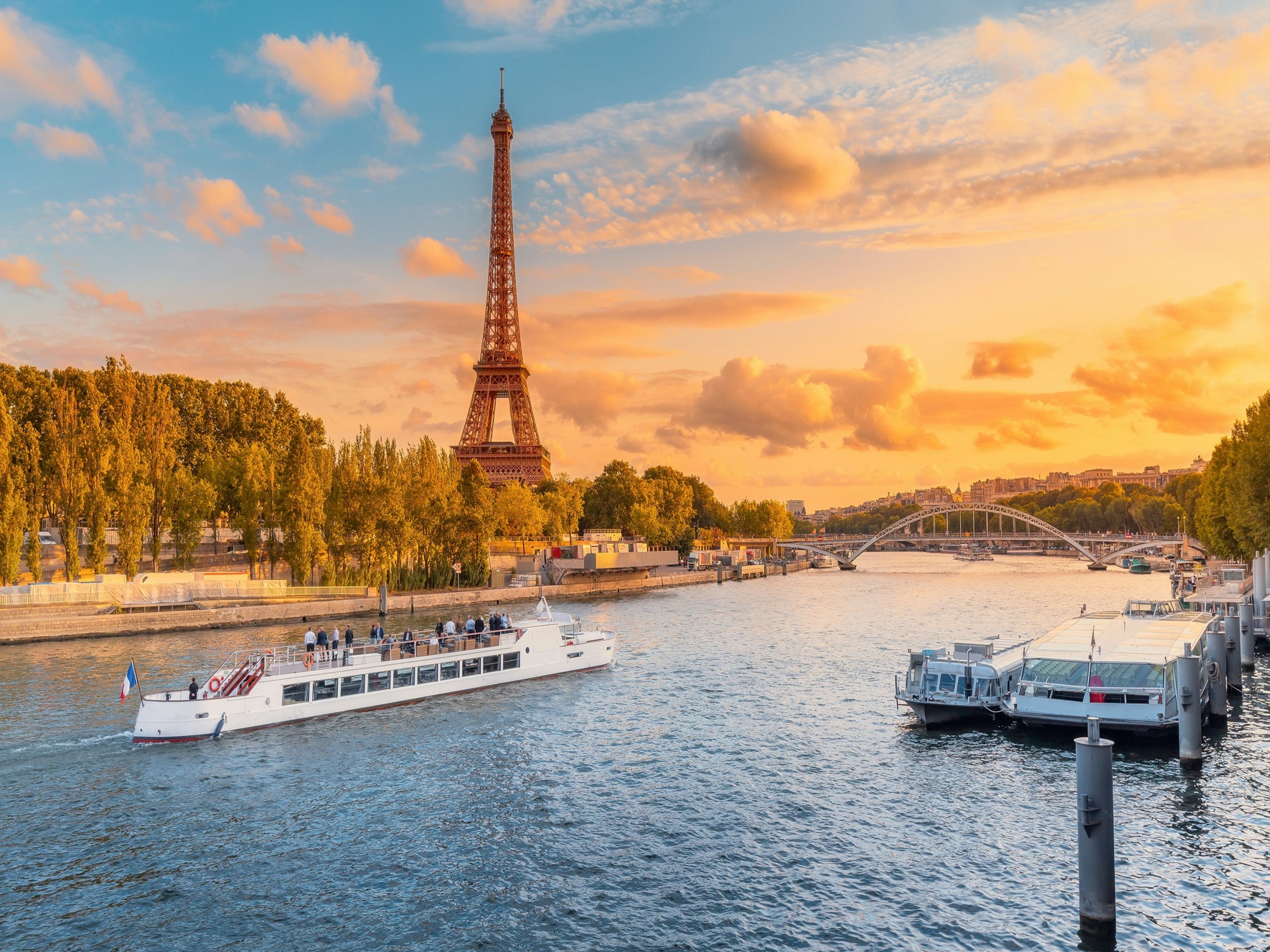 The image captures the iconic Eiffel Tower at with an orange hued sky at dusk with a tourist boat on the Seine River in Paris.