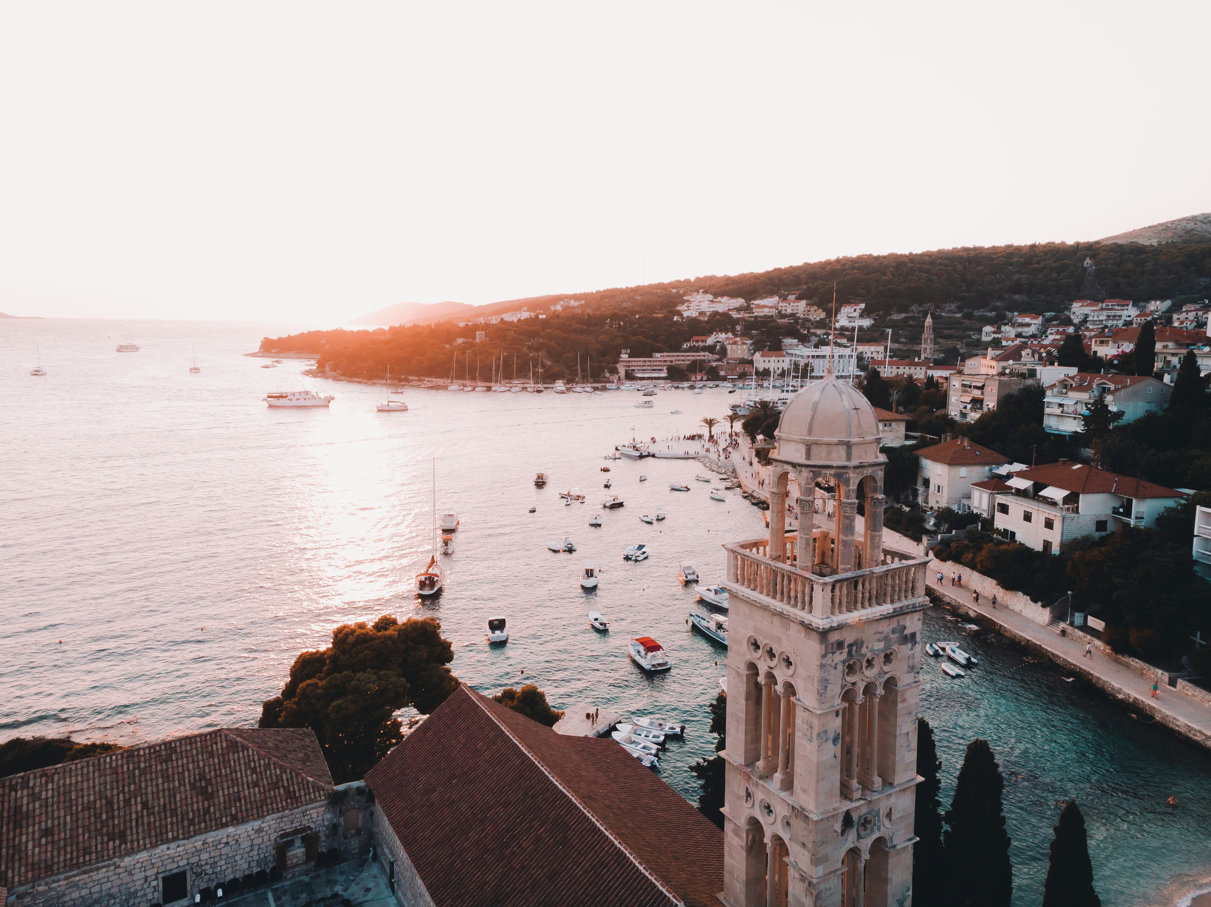 A beautiful view of the coastline of Hvar with a tall bell tower and many boats on the ocean and hills splattered with buildings.