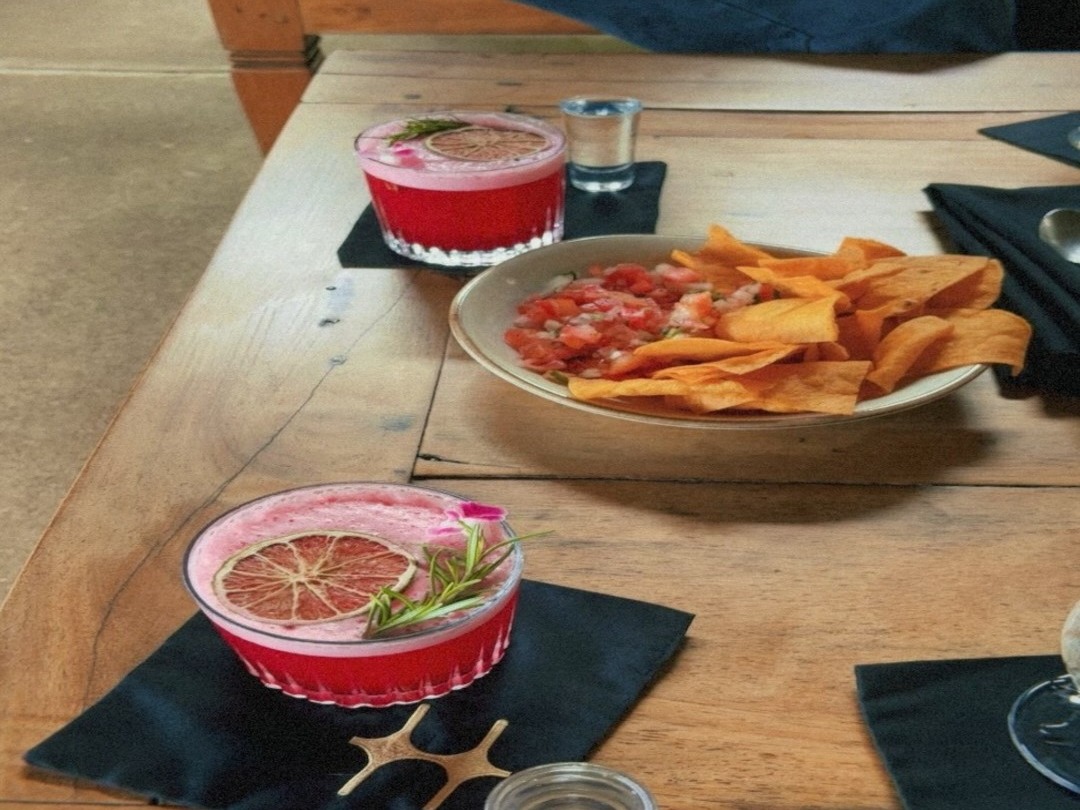 The image features a table with two pink drinks garnished with lime, a bowl of nachos with salsa, and a person in the background.