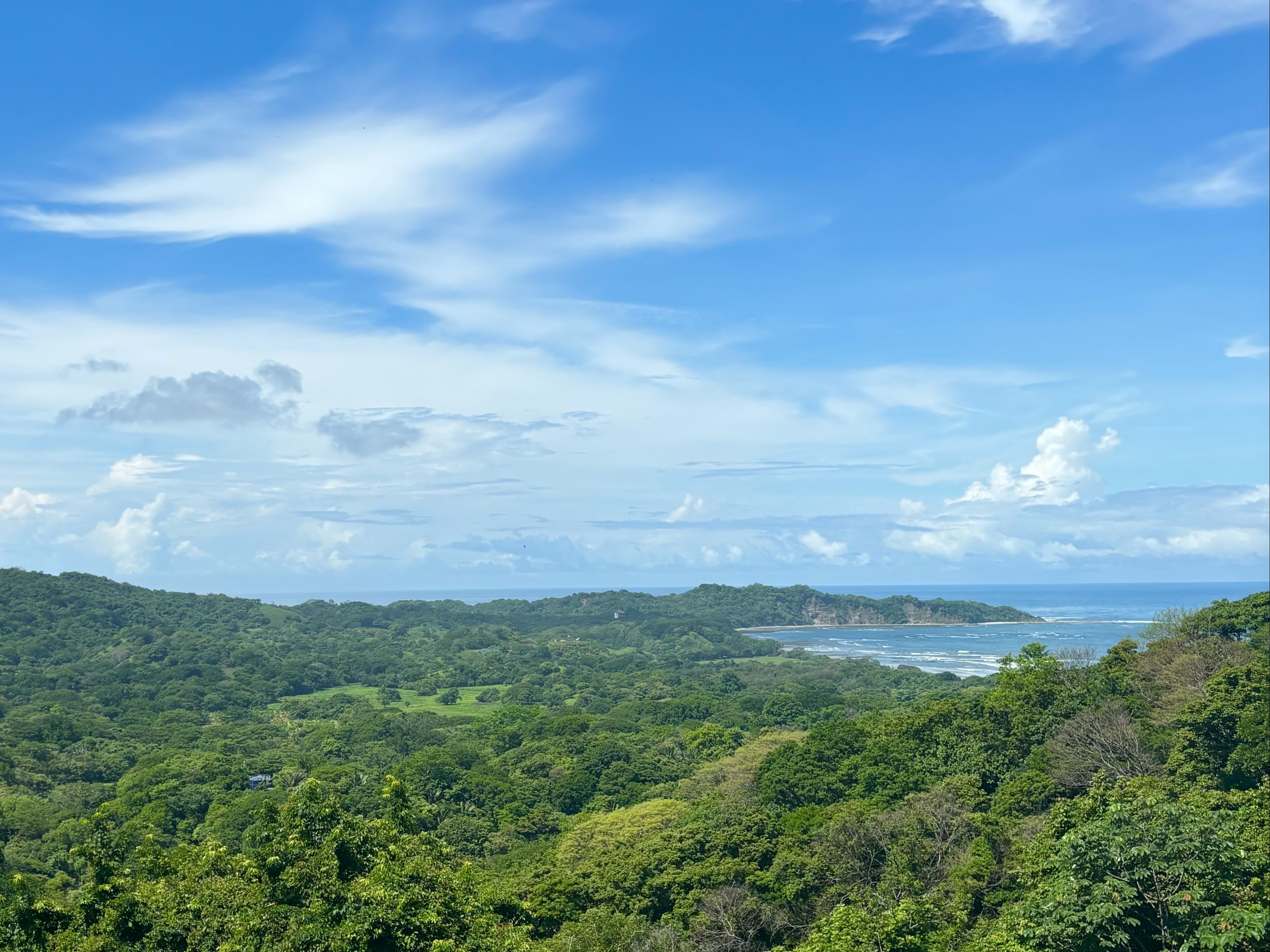 The image displays a lush green landscape with dense foliage and a glimpse of the ocean in the distance under a clear blue sky with scattered clouds.