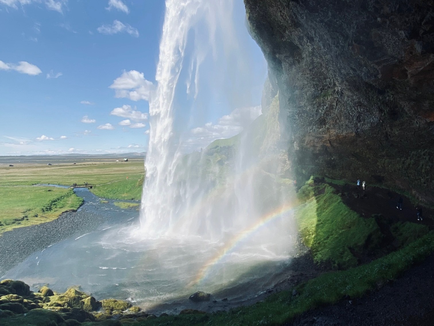 A small rainbow formed by a waterfall with a flat grassy plane in the distance on a sunny day.