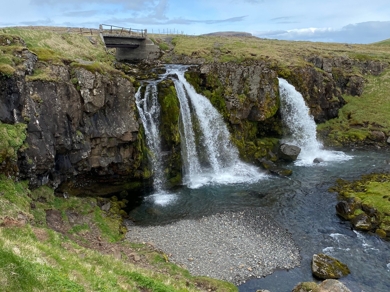 Three waterfalls with a cliff descending into a body of water surrounded by greenery on a sunny day.