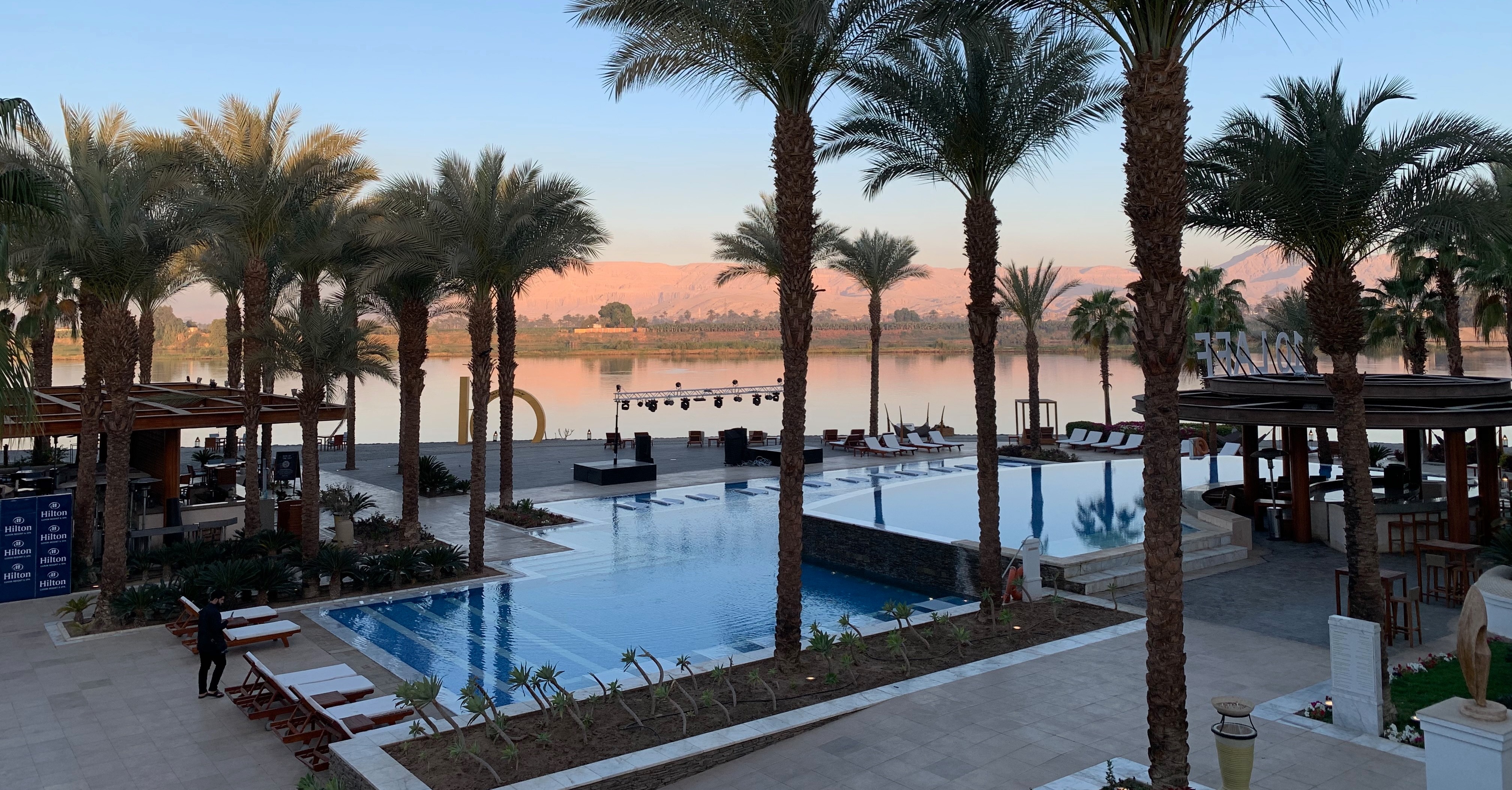 The image portrays a serene resort pool area with palm trees, beside a calm river and the desert in the distance.